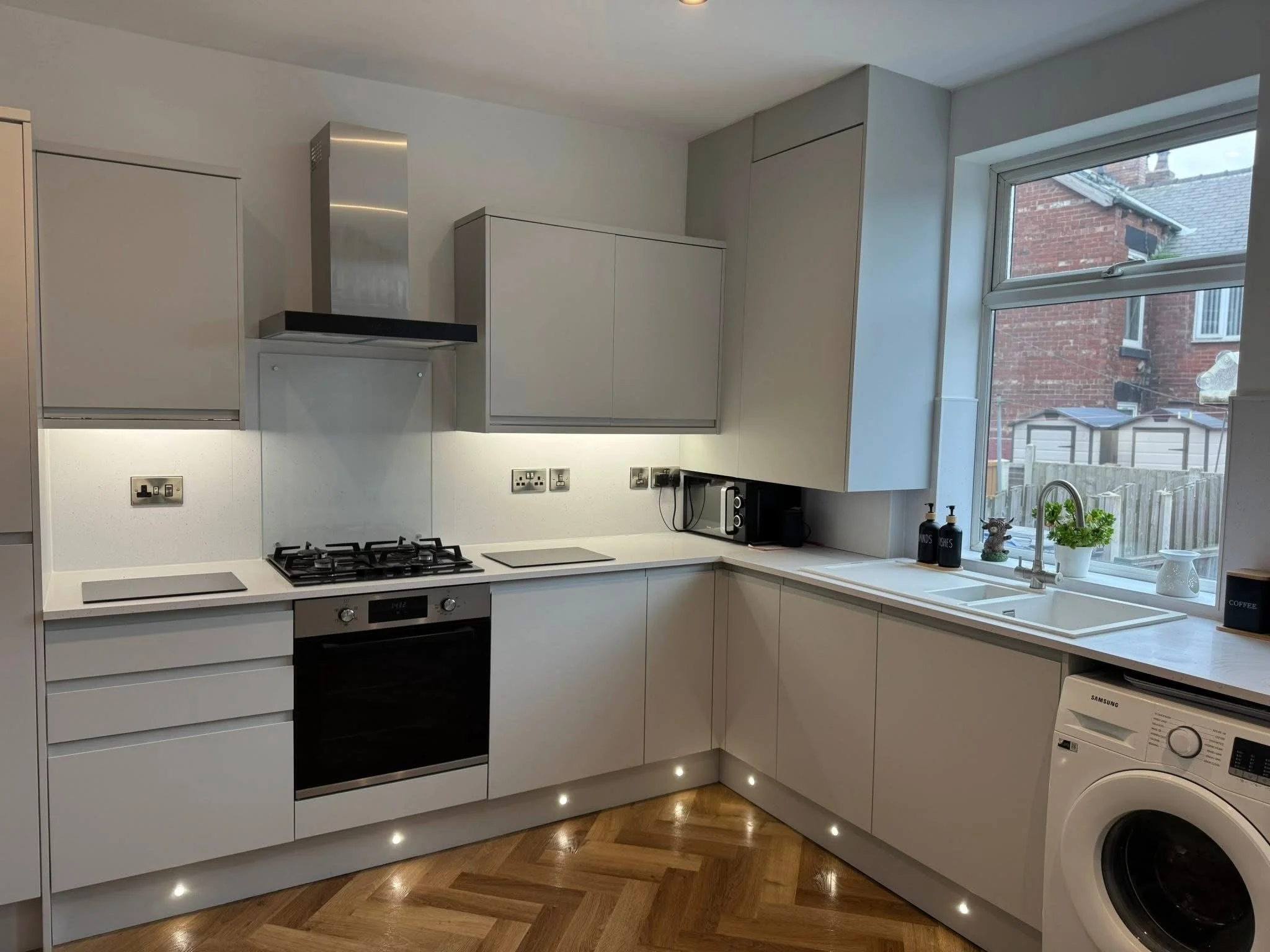 Modern kitchen with white cabinets, built-in oven, black stovetop, and large window showing neighboring brick houses.
