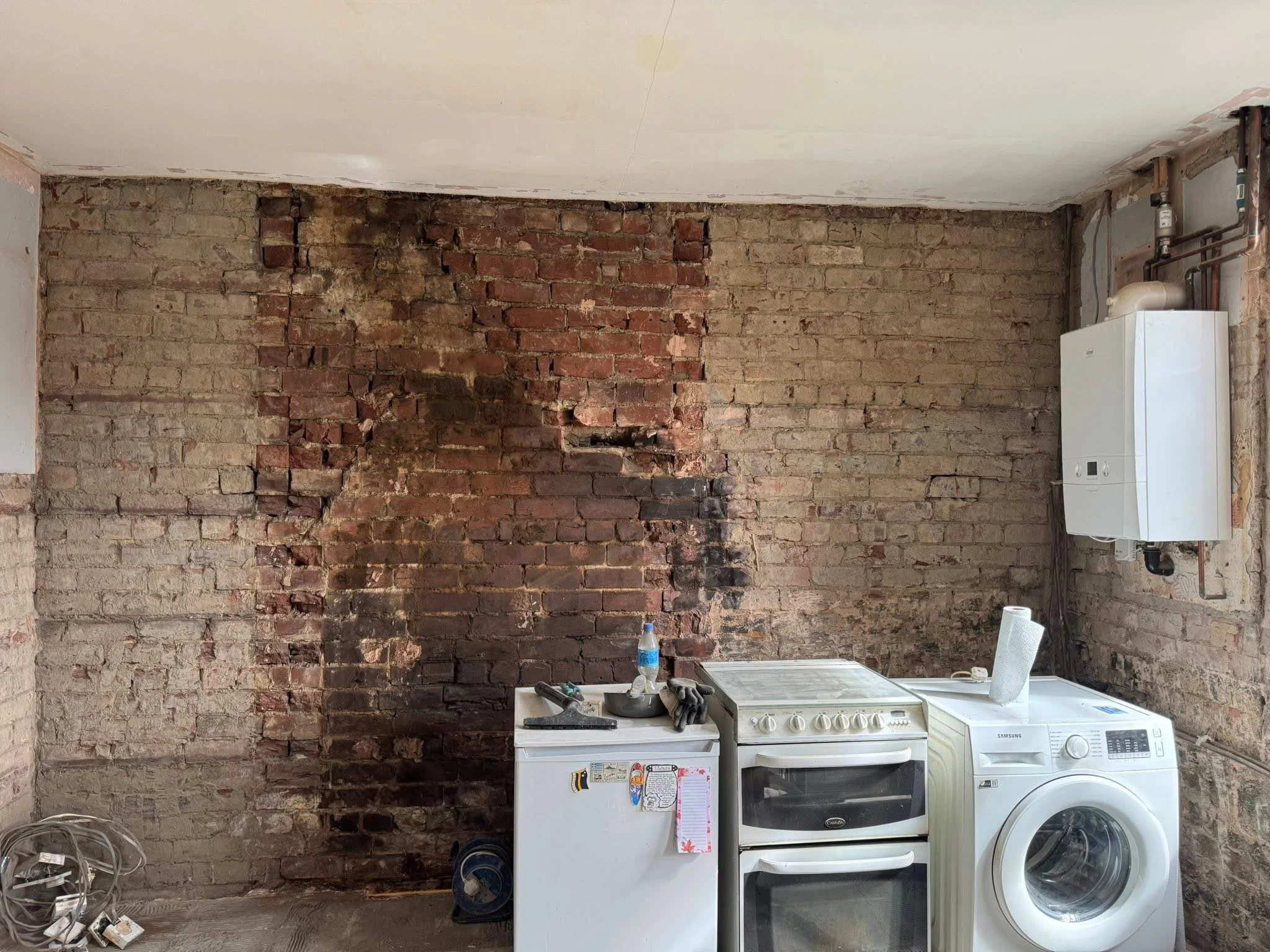 Room with exposed brick wall, white ceiling, and laundry appliances including a washing machine and a dryer. Items on top of the dryer include a bottle of water, a gray bowl, and gloves. The wall has some patches of darker bricks and black soot or smoke stains.