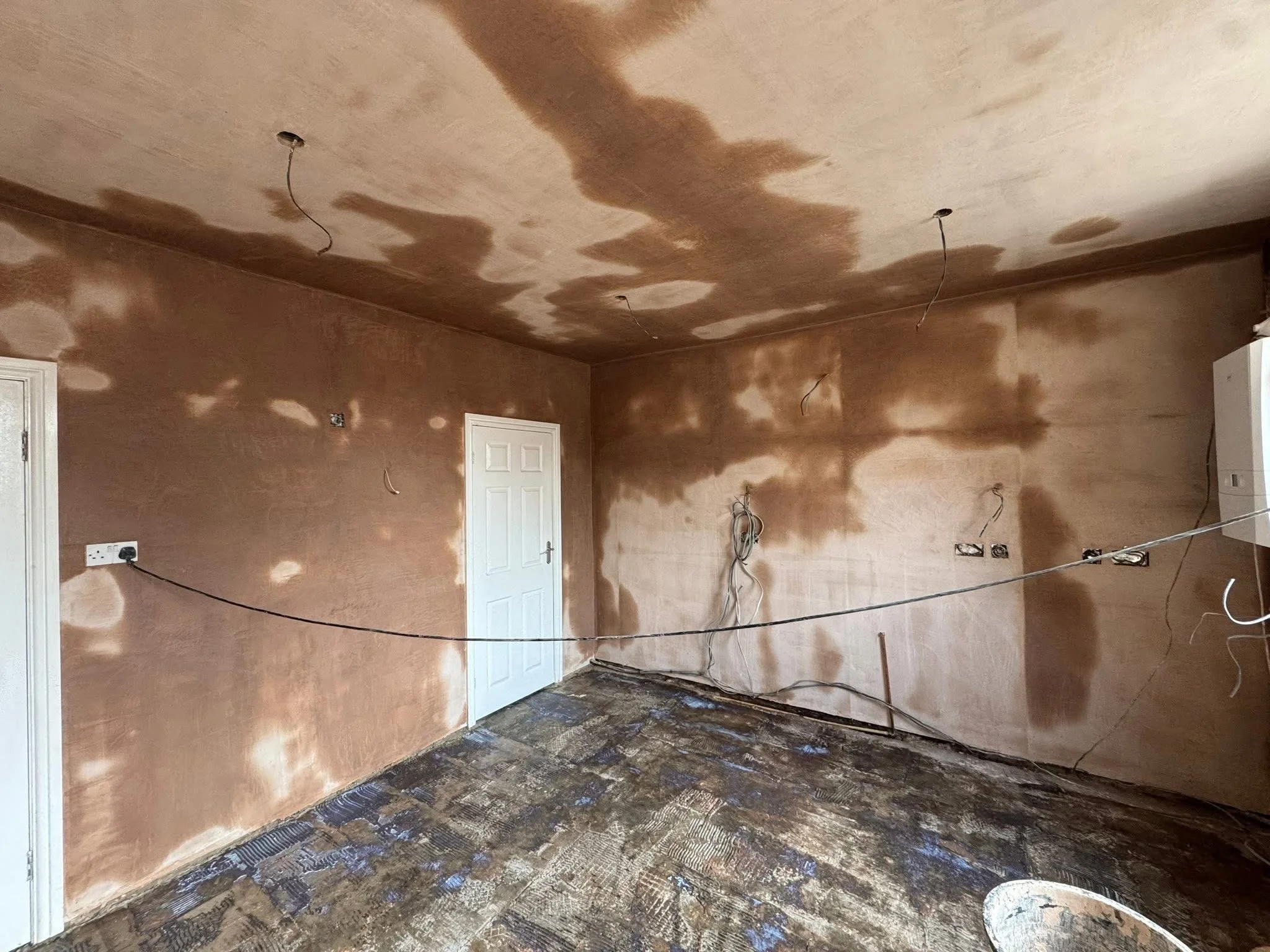 A room under renovation with brown walls and ceiling, electrical wires hanging from the ceiling, and a partially removed flooring revealing subflooring.