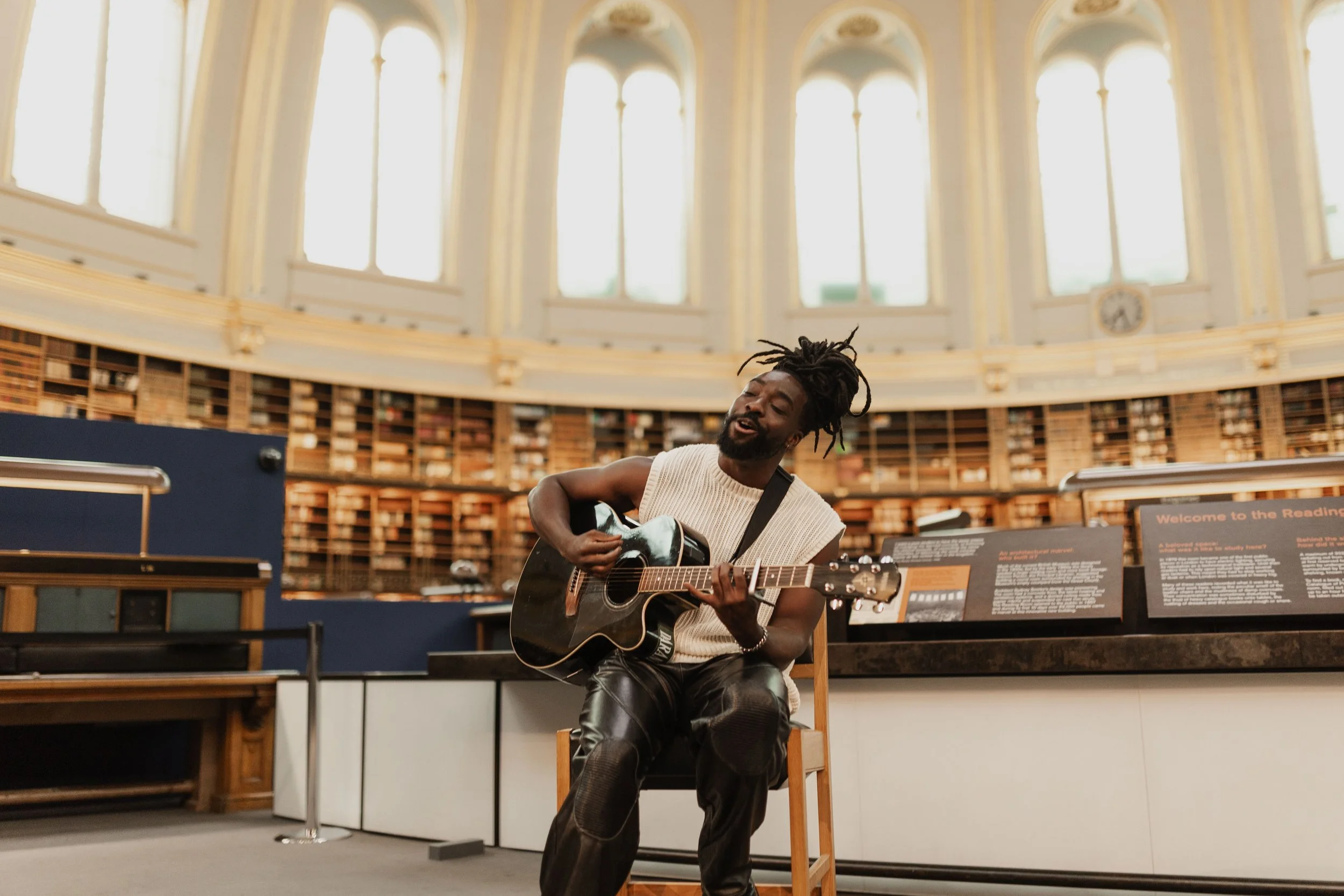 A man with dreadlocks playing acoustic guitar and singing in a grand library with tall arched windows and shelves full of books.