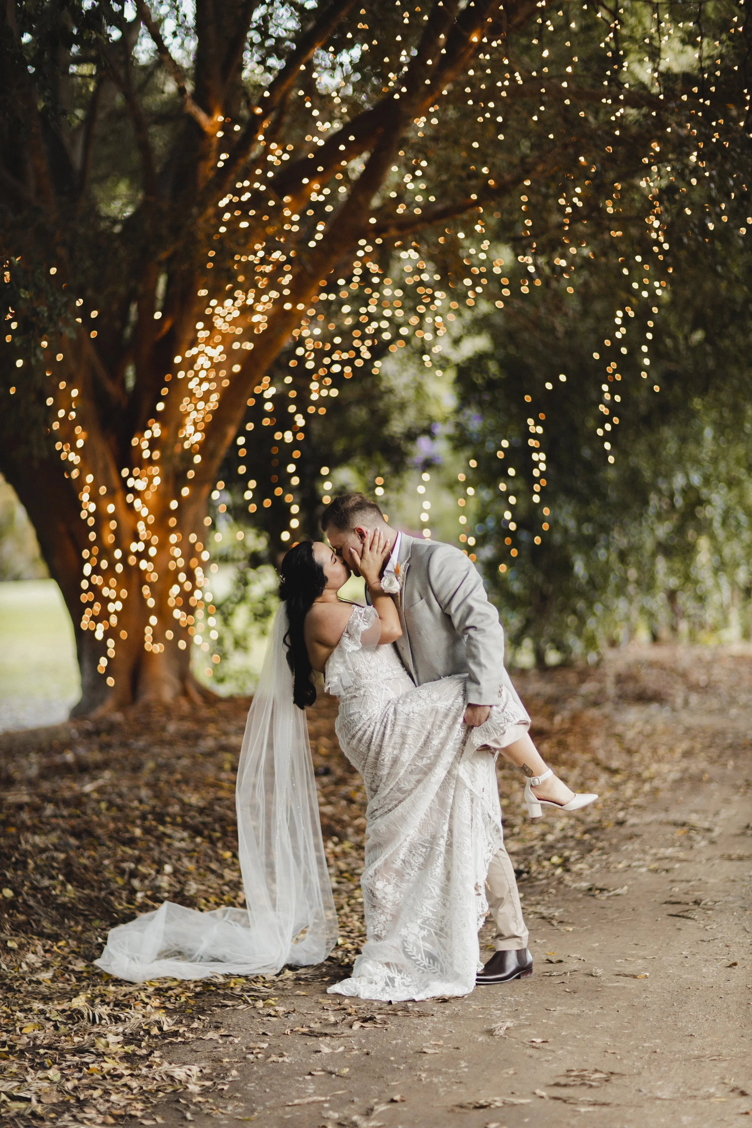 A newlywed couple sharing a romantic kiss under a large tree decorated with string lights, with the groom holding the bride up as she wears a lace wedding dress and veil, and the groom in a light gray suit, surrounded by autumn leaves on the ground.