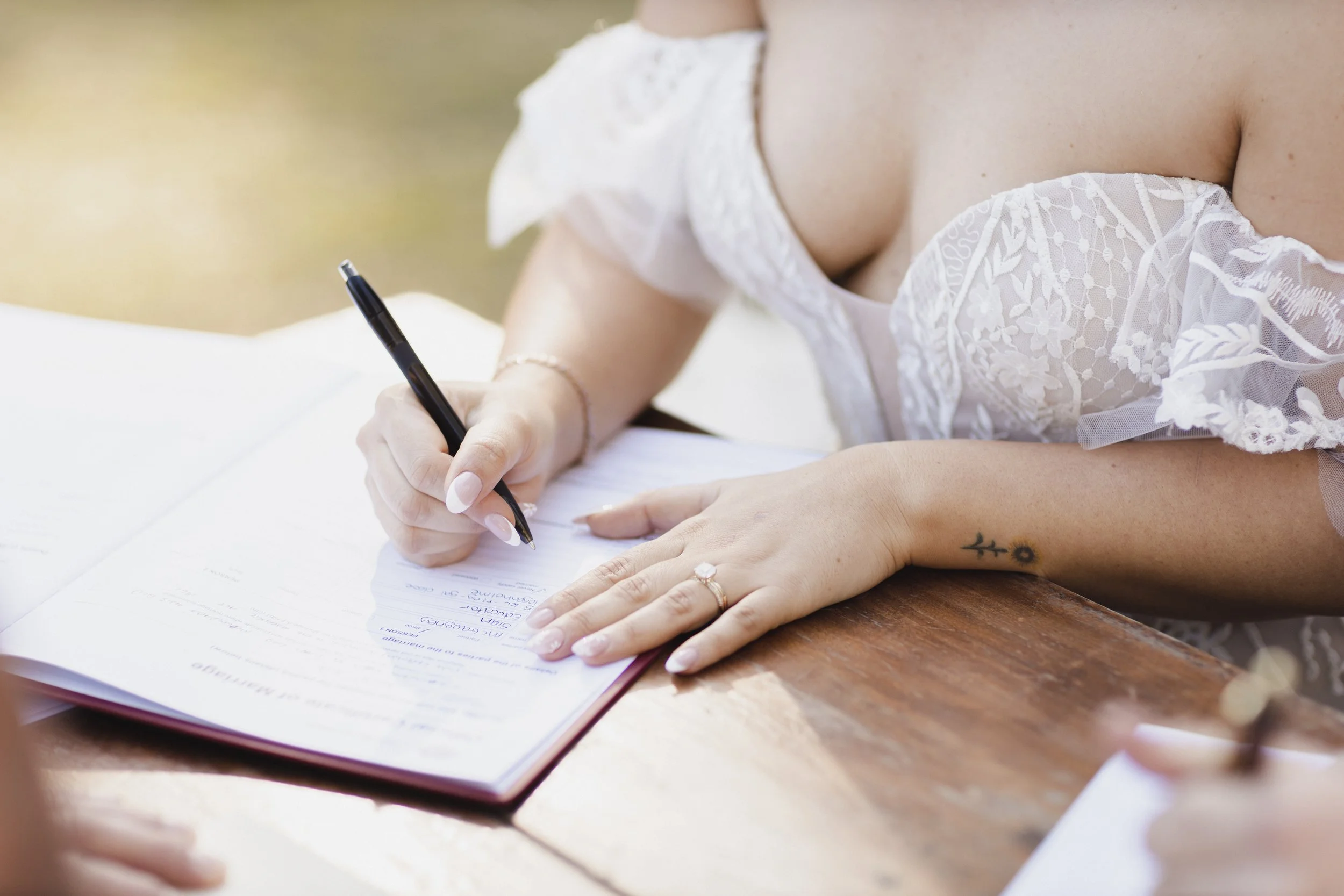 A woman wearing a white off-shoulder lace dress is signing a document on a wooden table with a black pen. She has a small tattoo of a branch with leaves on her forearm and a ring on her finger.