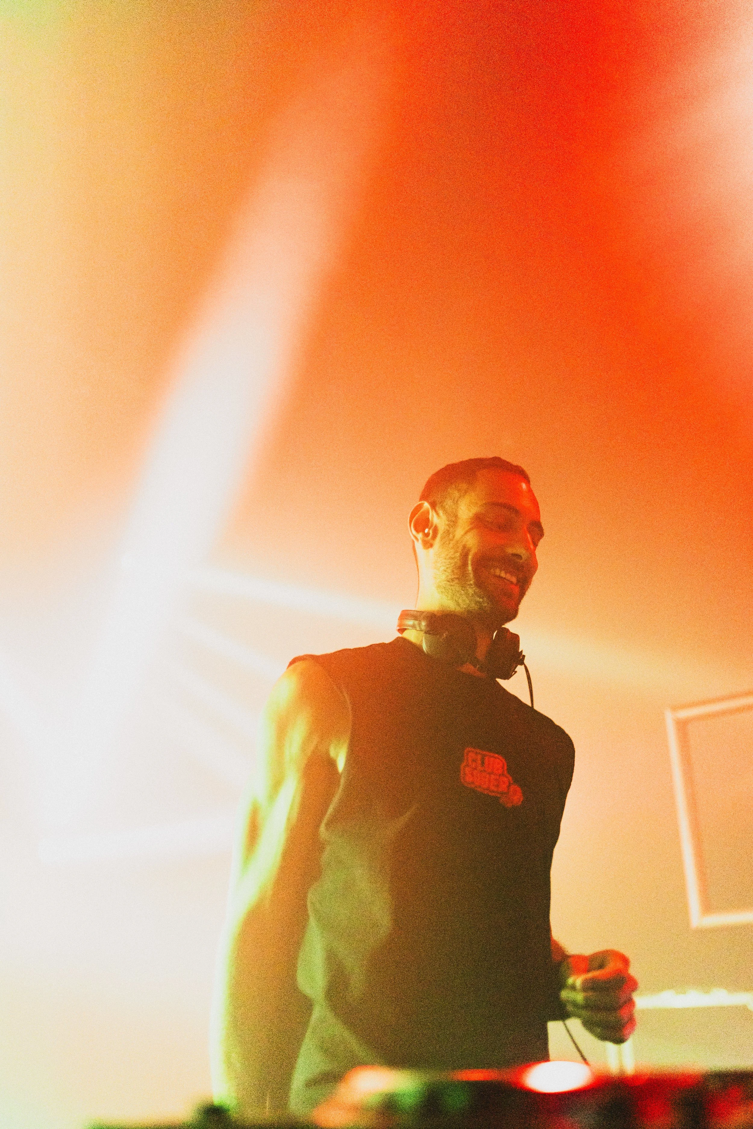A DJ with headphones around his neck smiling at a lively party scene with orange lighting.