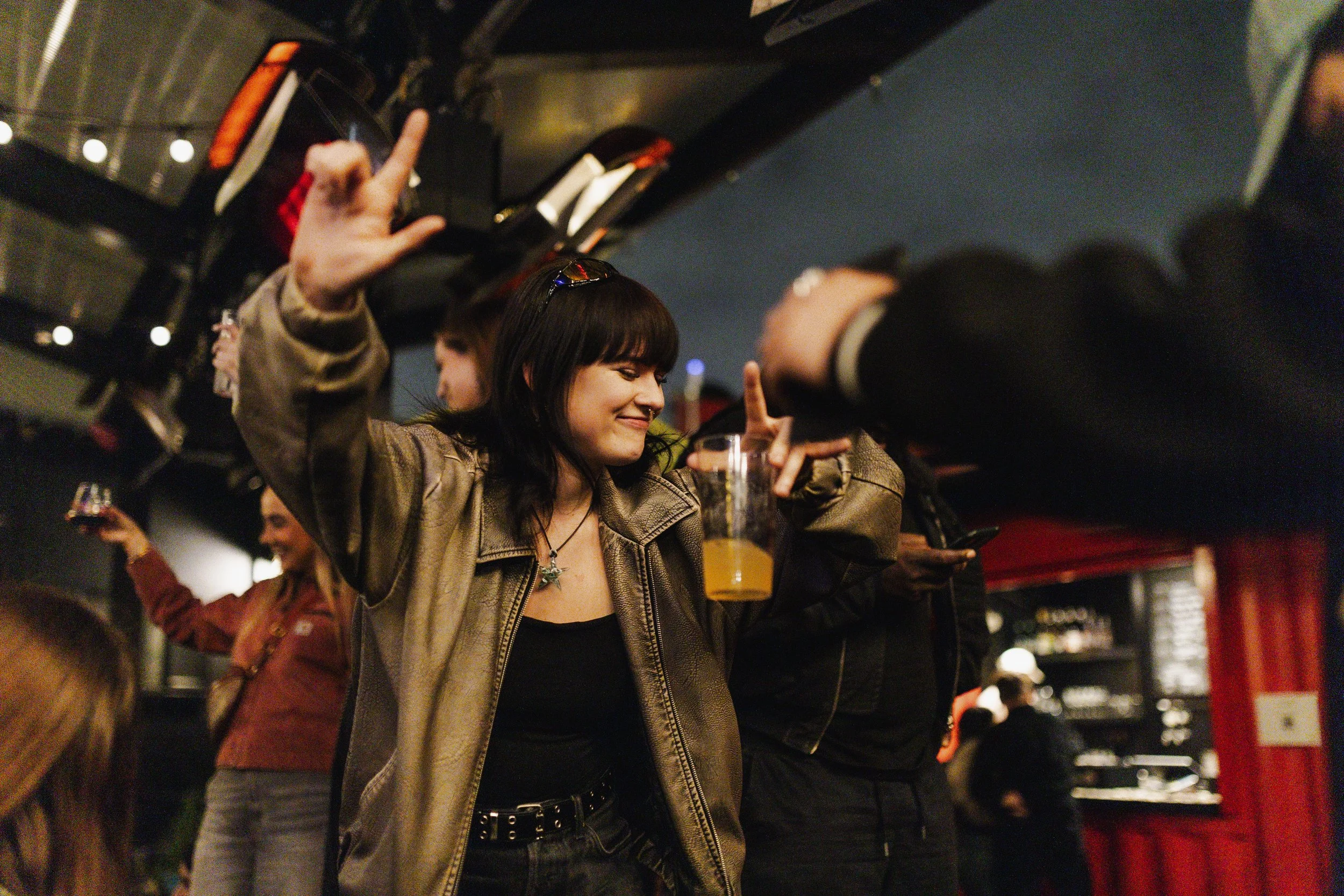 A woman in a leather jacket dancing and smiling at a party, holding a drink, with other people in the background.