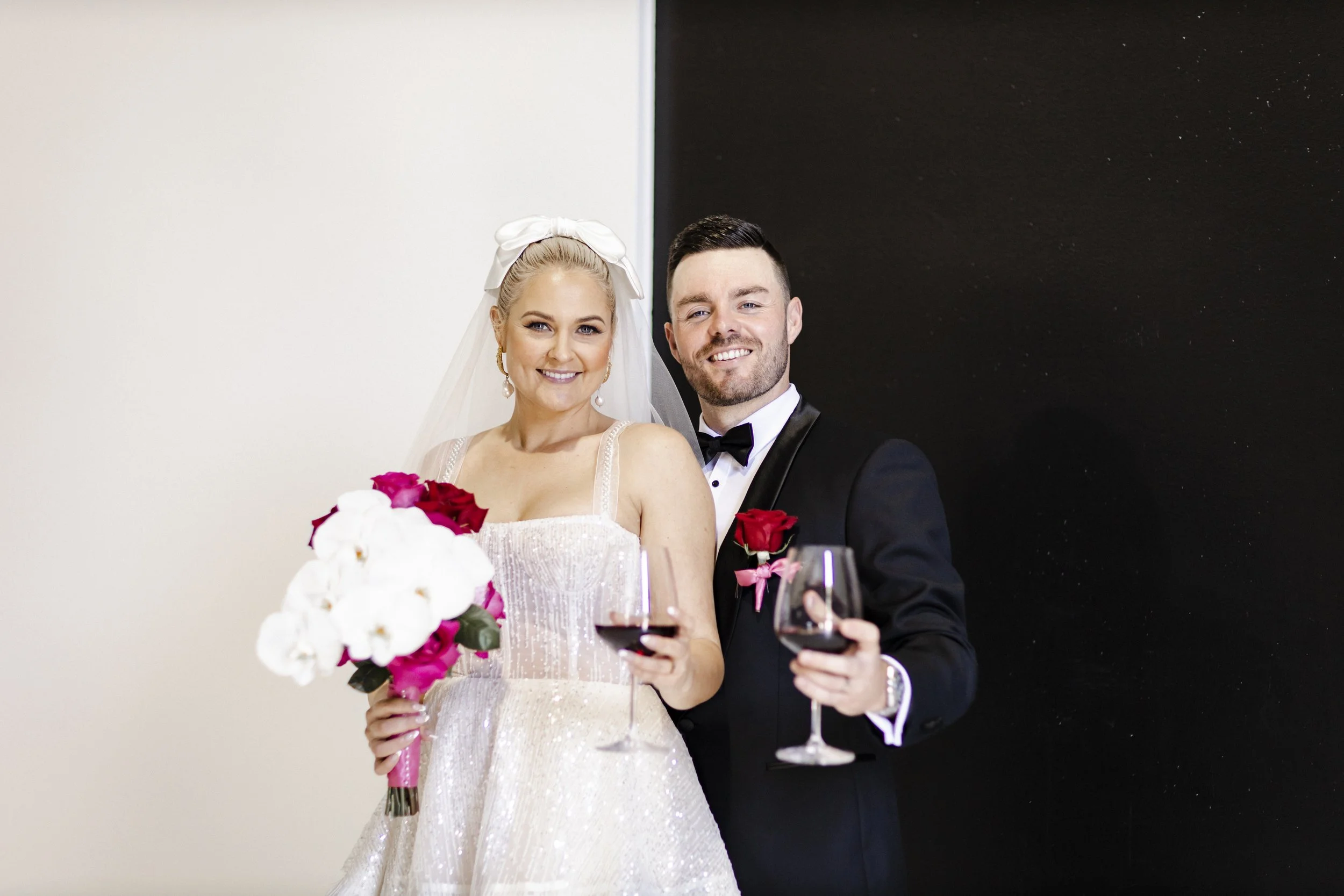 Bride and groom in wedding attire holding glasses of red wine and smiling at the camera.