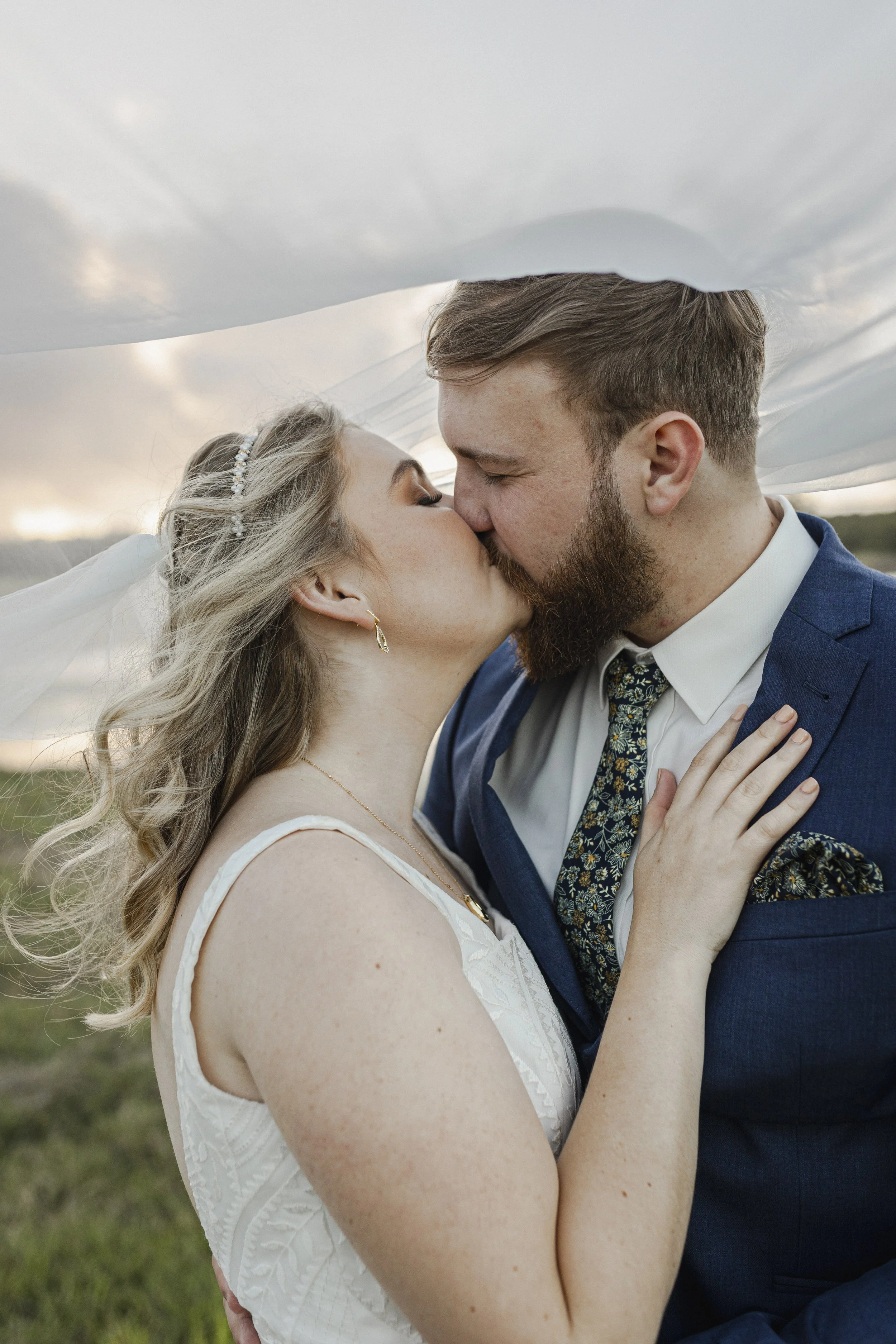 A bride and groom sharing a kiss outdoors, with a veil overhead and a cloudy sky in the background.
