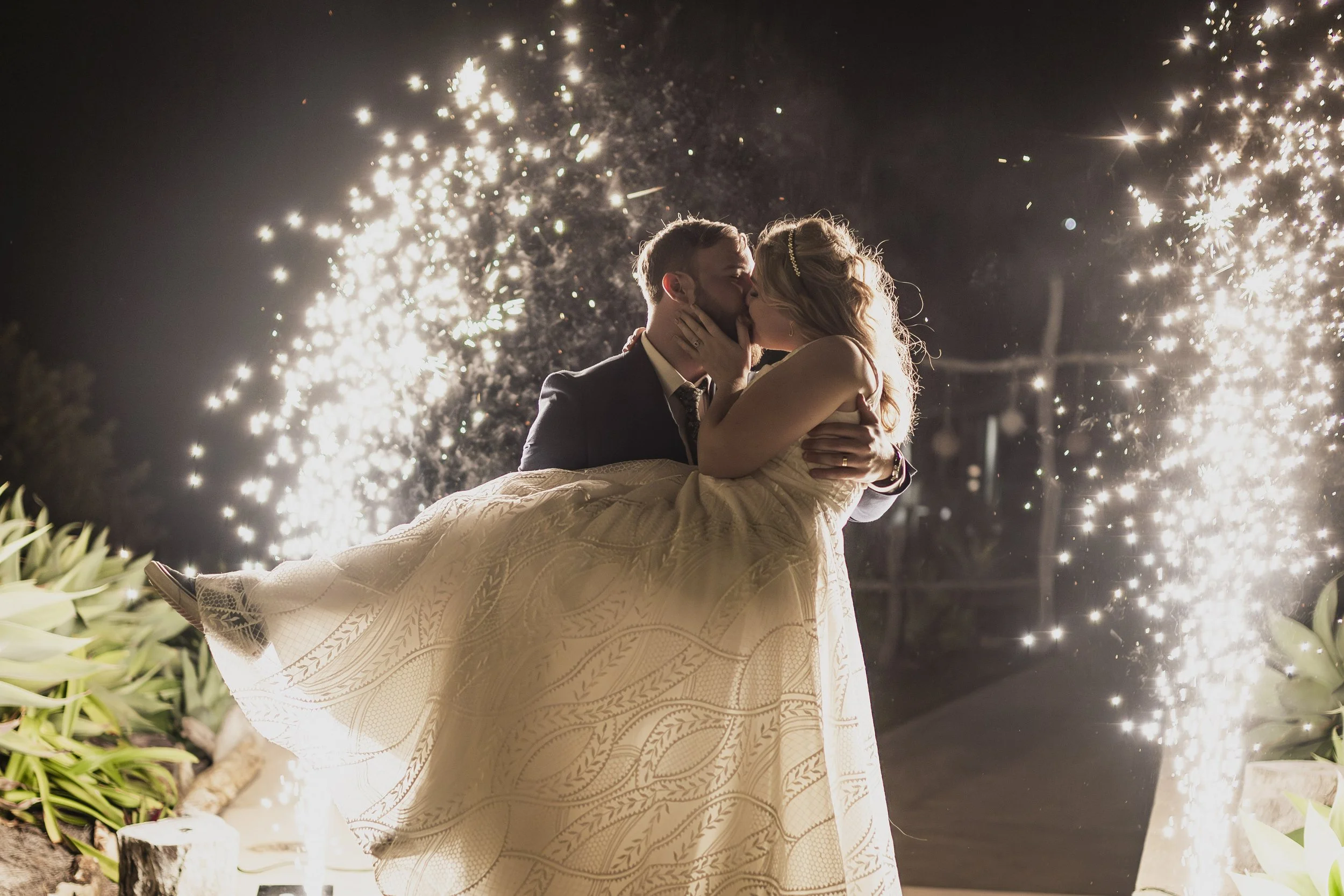 A newlywed couple sharing a kiss at night while the groom carries the bride, with sparklers creating a bright display in the background.