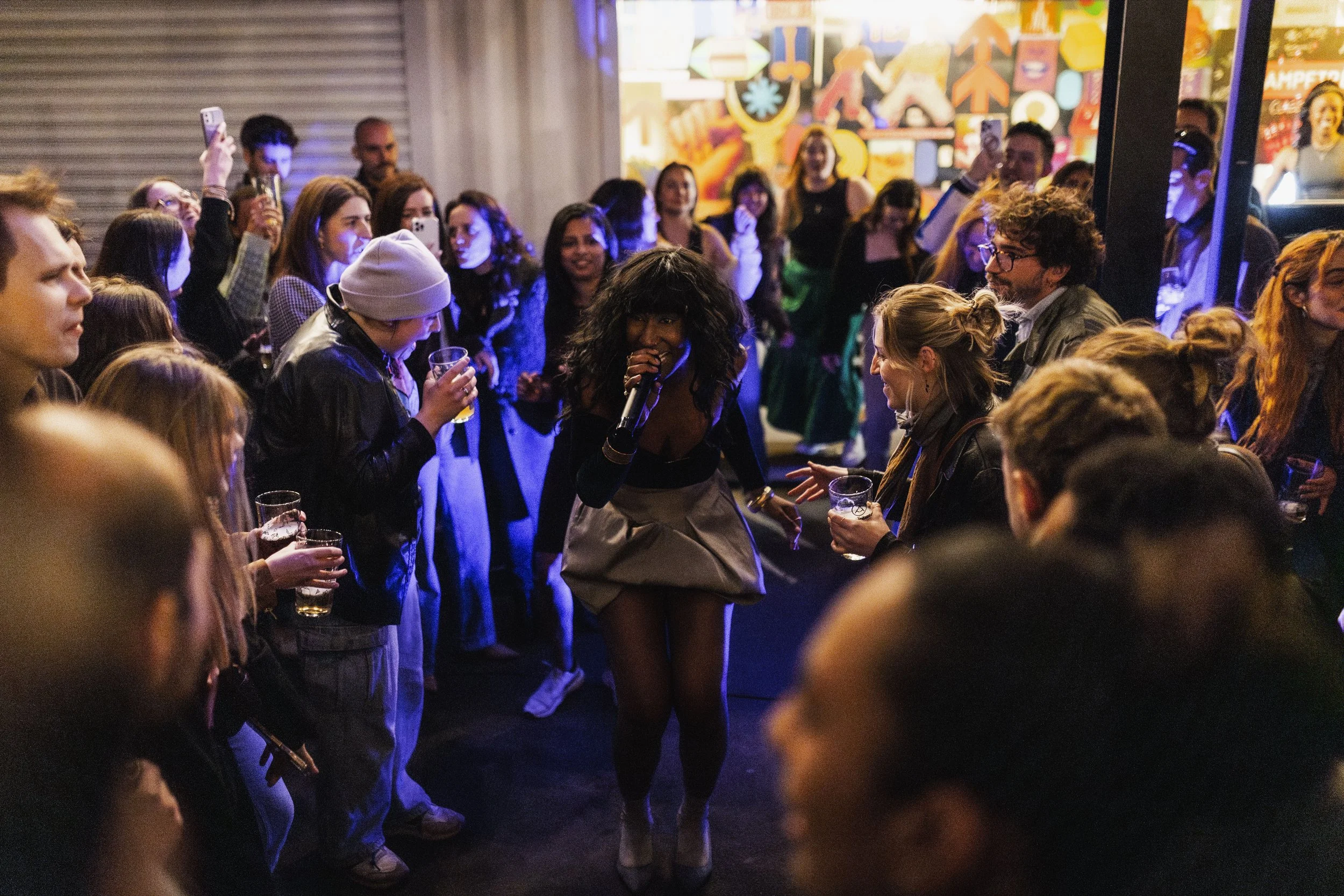 A woman with curly black hair, wearing an elegant dress, is singing into a microphone at a lively party where guests are dancing and holding drinks.