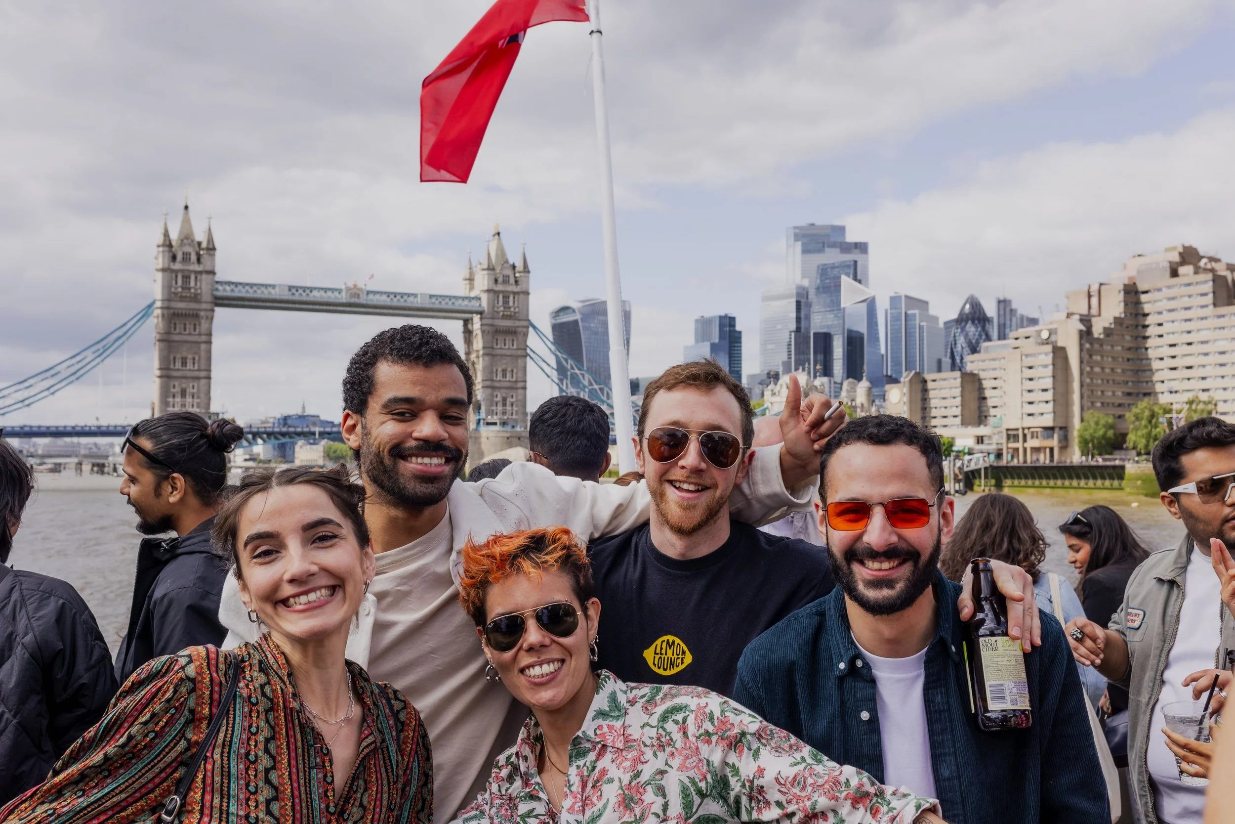 Group of smiling friends posing outdoors near Tower Bridge in London with the city skyline in the background.