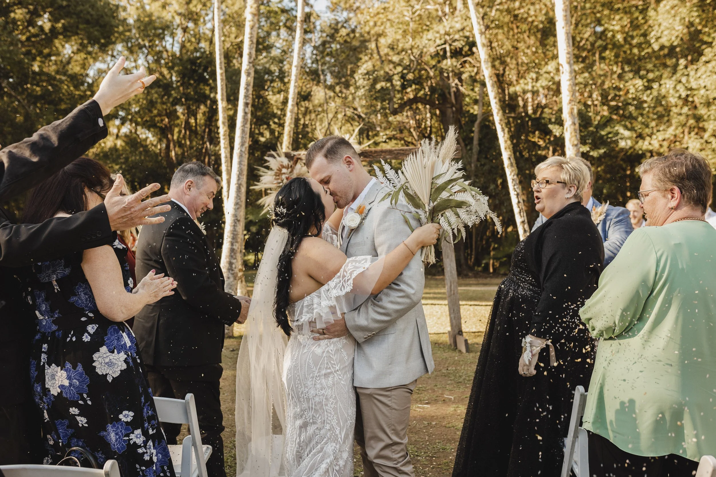 A bride and groom kiss during their outdoor wedding ceremony, surrounded by friends and family throwing confetti.