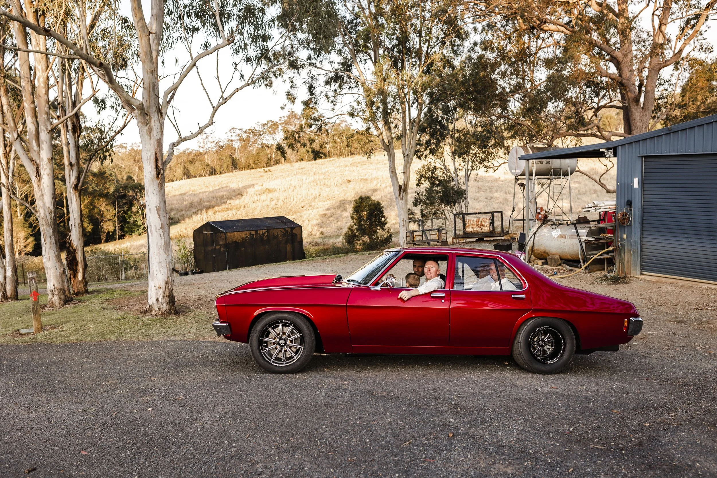 A red vintage car with three people inside, parked outdoors near trees and a fenced area, during sunset.