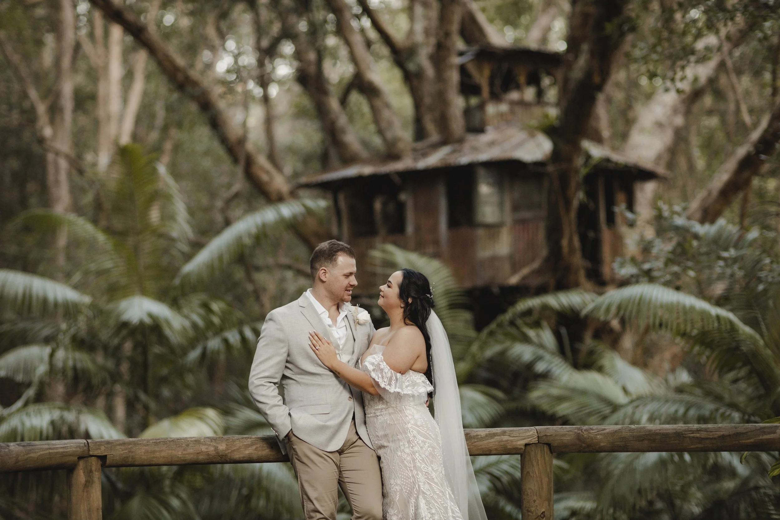 A newlywed couple standing on a wooden bridge in a forest, gazing at each other. The bride is wearing a white lace wedding dress and the groom is in a light gray suit. In the background, there is an old treehouse among the trees, surrounded by lush g