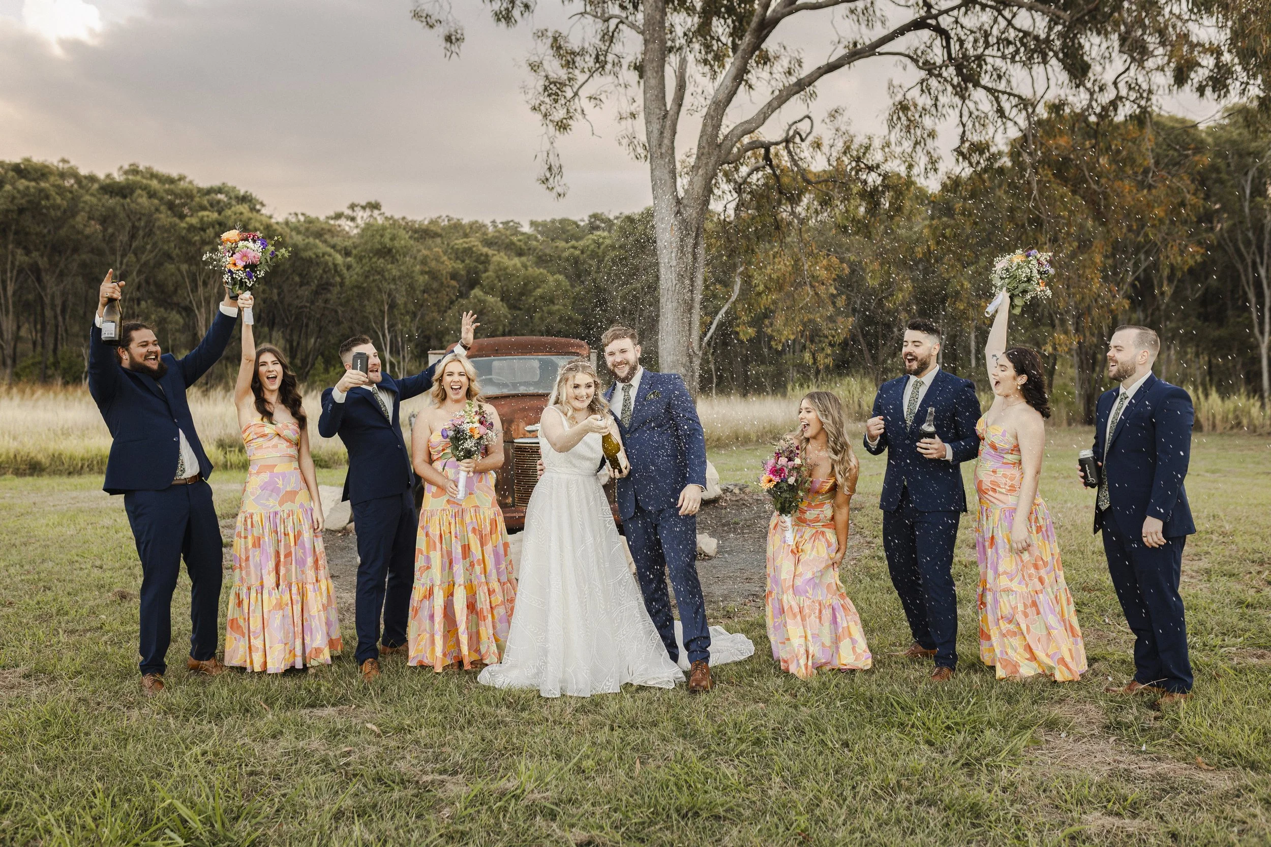 A wedding celebration outdoors showing a bride and groom surrounded by bridal party members holding bouquets and drinks, with some spraying champagne and celebrating in an open grassy area with trees and an old truck in the background.