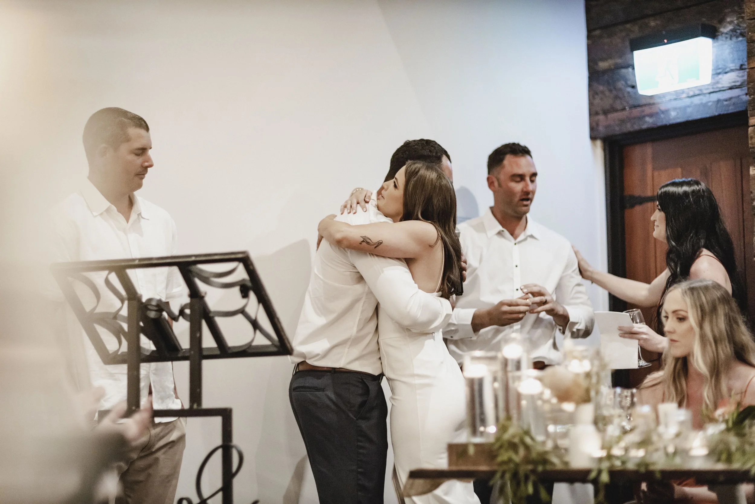 A group of people at a wedding reception, with two women hugging in the center, surrounded by friends and family.