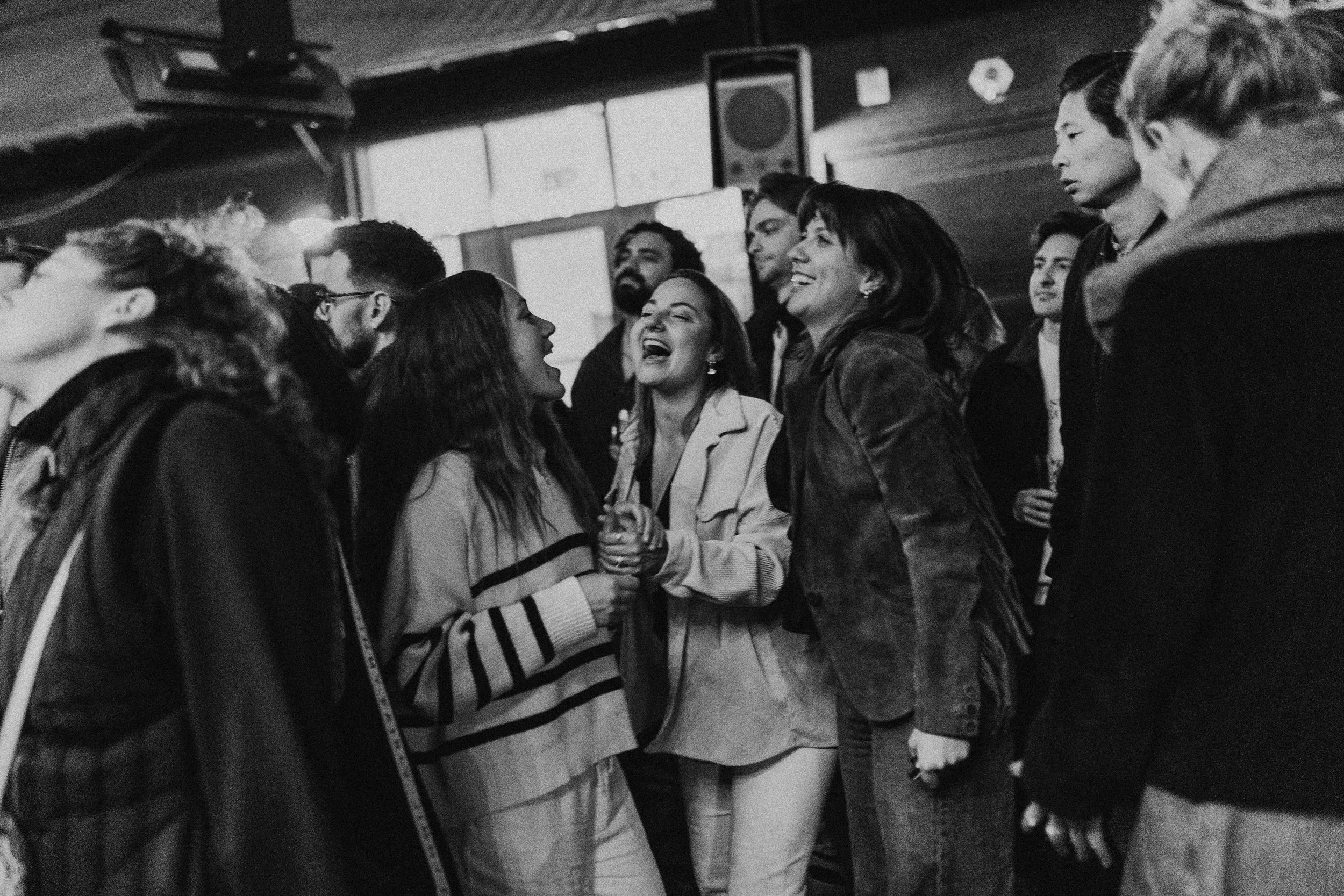 Black and white photo of a group of people at a social gathering, with two women in the center holding hands and smiling, engaging in lively conversation, surrounded by other individuals.