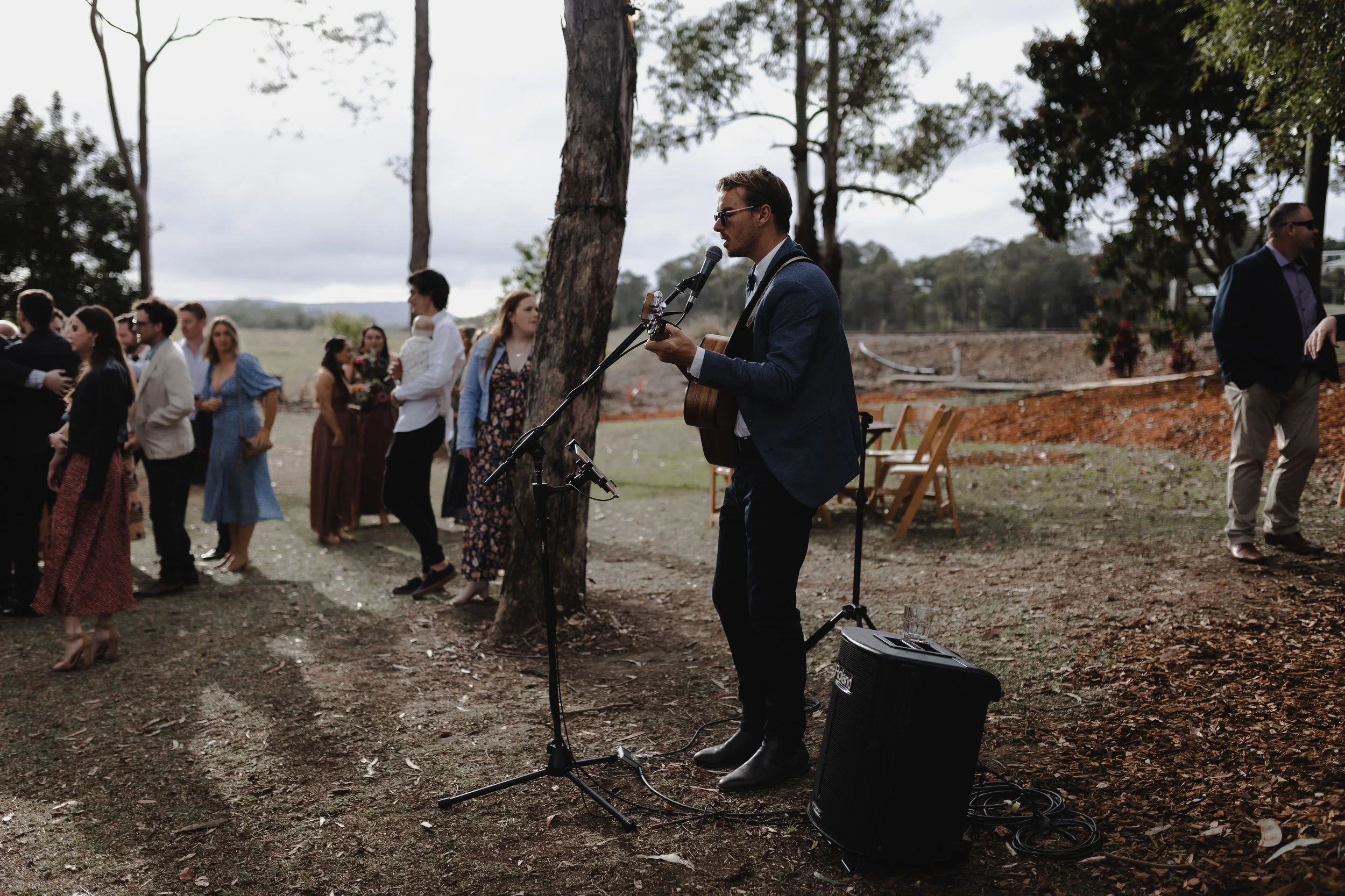 A man in a blue jacket playing guitar and singing into a microphone outdoors, with a group of people standing in line behind him, at a rustic event in a wooded area.