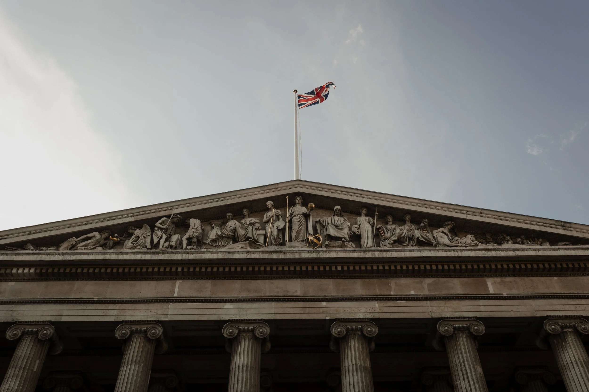 The upper part of a classical building with large columns and a detailed sculptural frieze featuring human figures. A Union Jack flag is flying atop a flagpole on the building's roof against a partly cloudy sky.