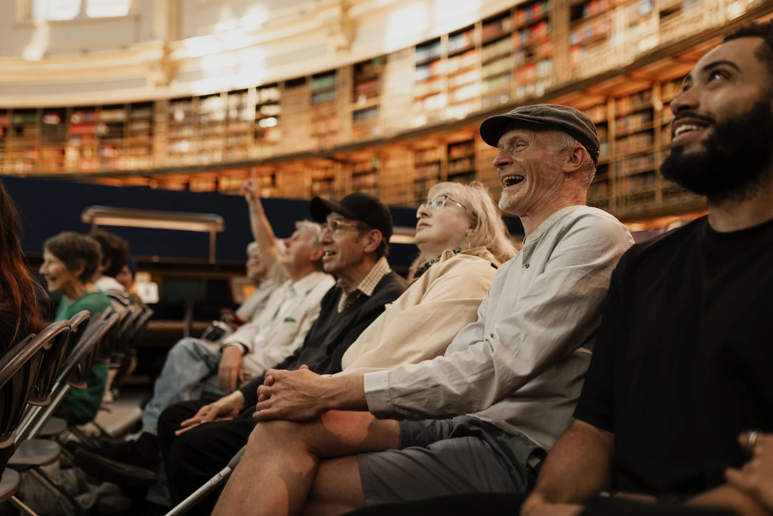 Crowd of people sitting in auditorium with bookshelves in the background, smiling and enjoying a lecture or performance.