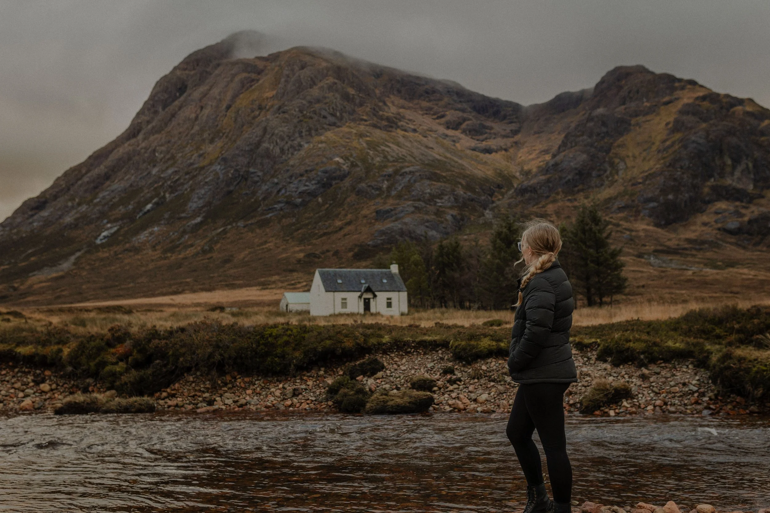 A woman with blonde hair in a braid, wearing a black jacket, stands in a river, looking towards a white house with a dark roof at the base of a mountain.