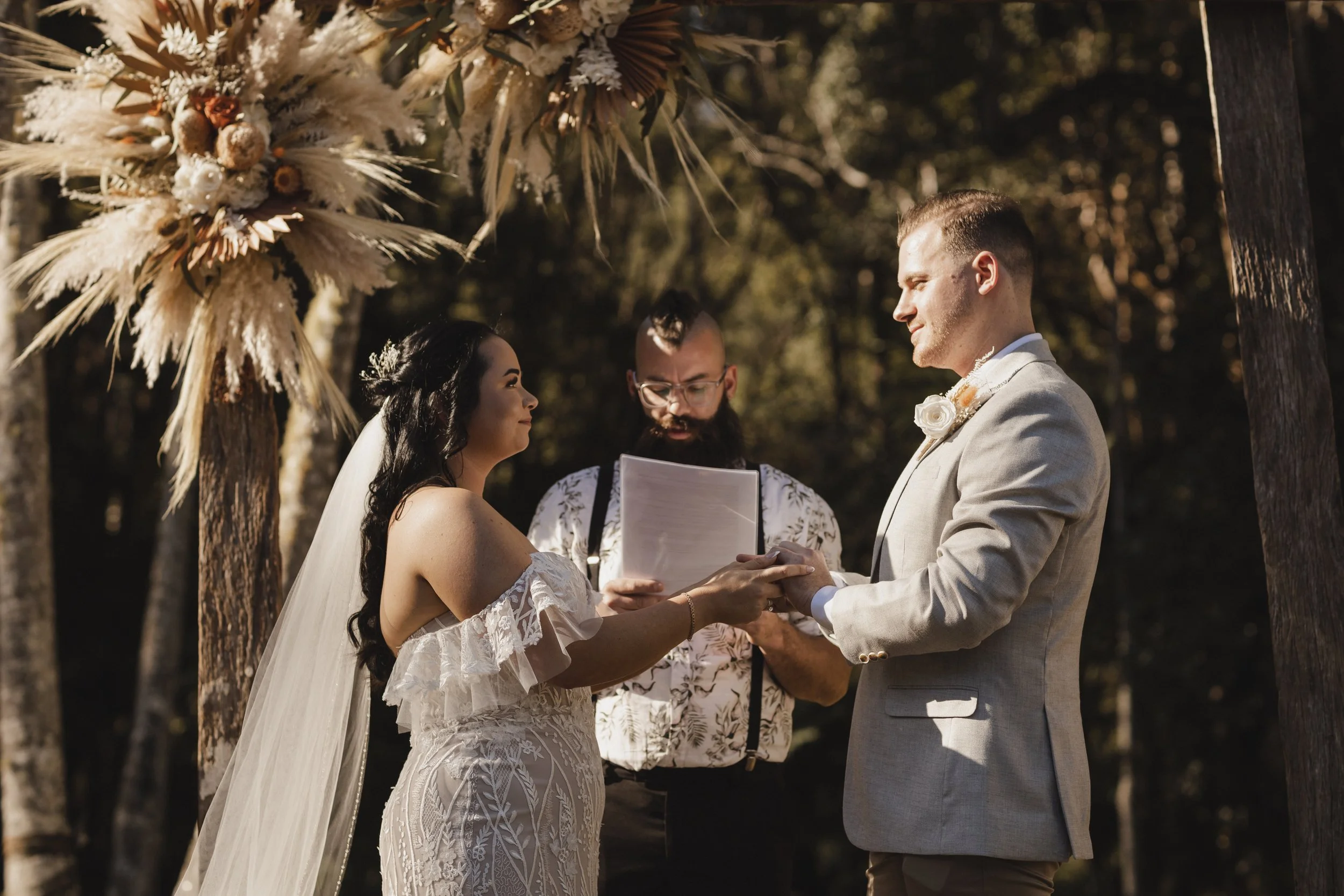 Bride and groom holding hands during outdoor wedding ceremony with officiant in background, under a floral arch in a wooded setting.