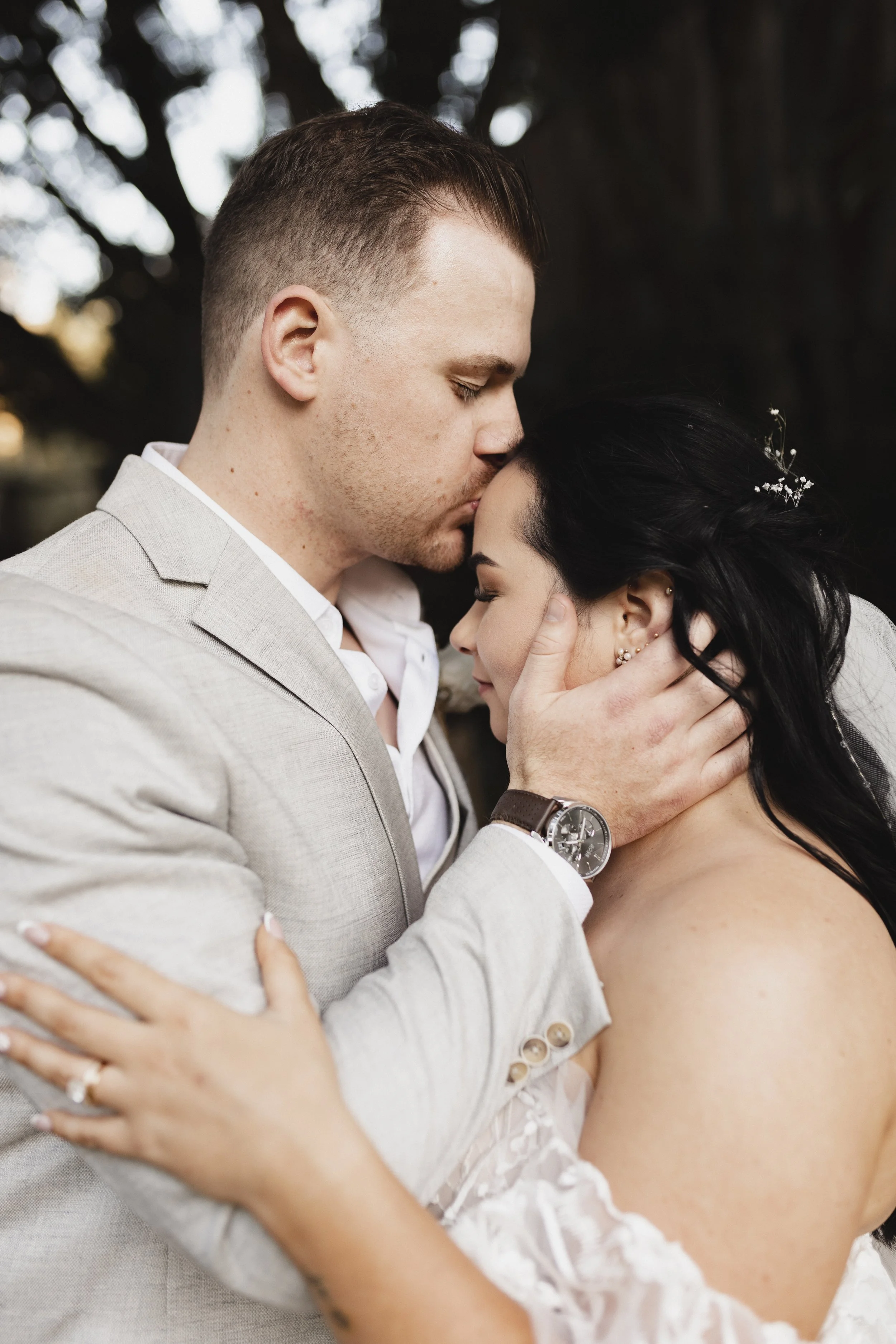 A man in a light-colored suit gently holding a woman with dark hair, wearing a wedding dress, as they share a tender moment outdoors.