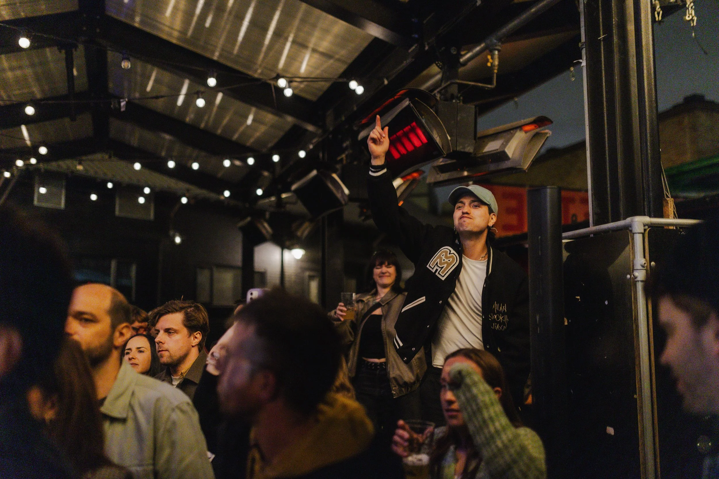 A young man standing with his arm raised, pointing upward, in a crowded indoor concert or event space with string lights and speakers hanging from the ceiling. Several people sit and stand around him, some holding drinks and taking photos.