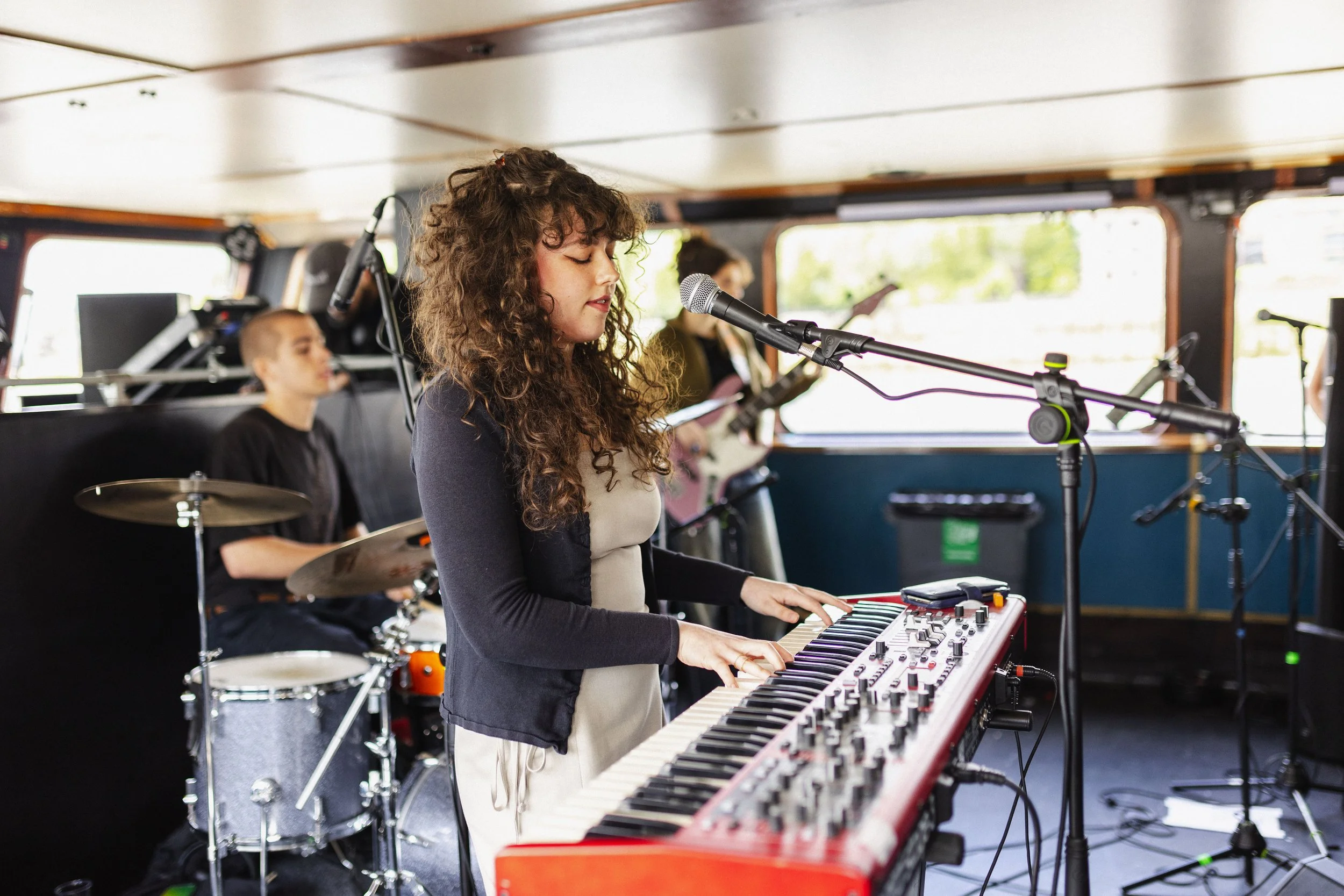 A young woman with curly hair singing into a microphone while playing a red keyboard, with two other musicians in the background—one on drums and another on guitar—in a room with large windows.
