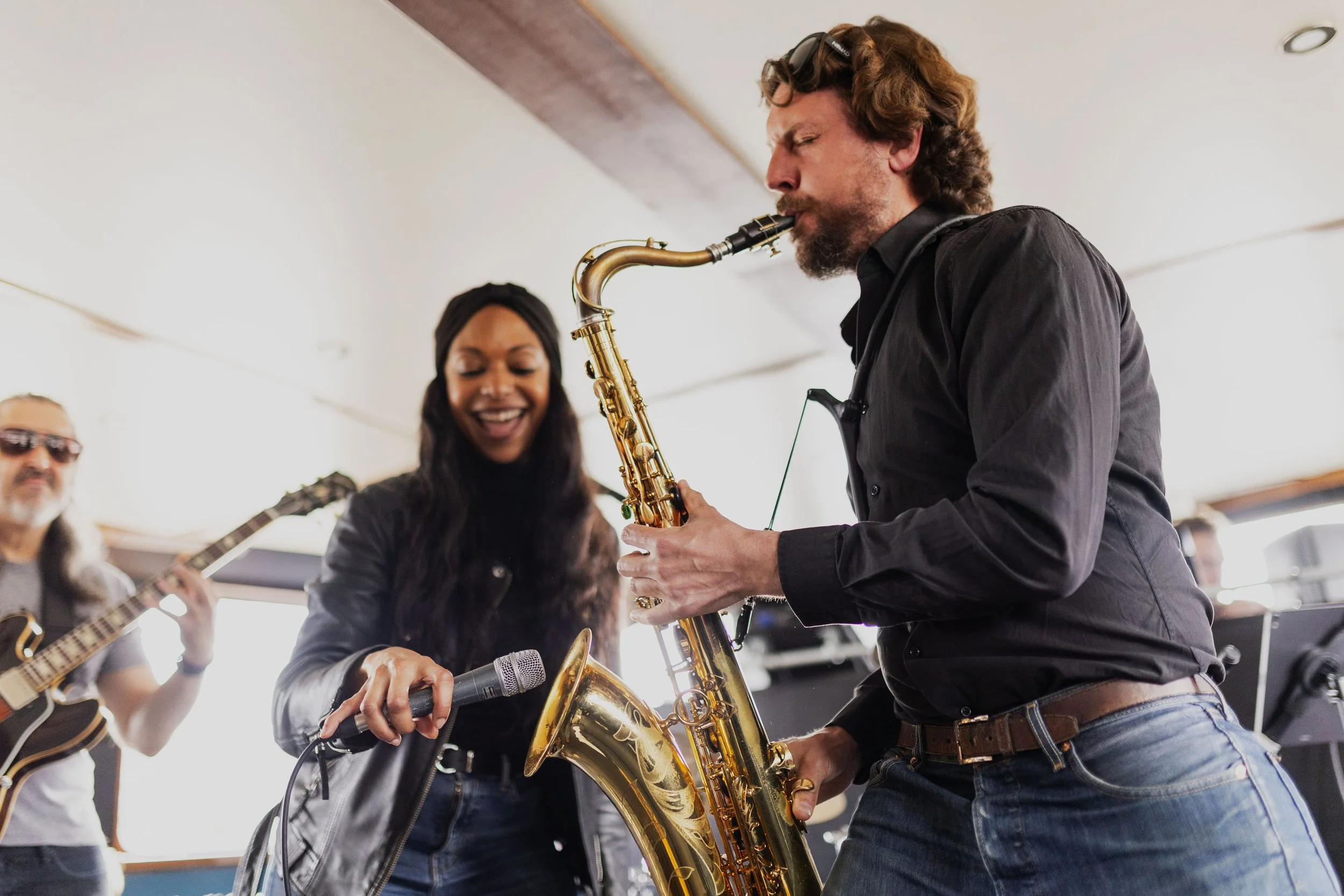 Musicians performing together in a bright indoor space, including a woman singing into a microphone and a man playing a saxophone, with a guitarist in the background.