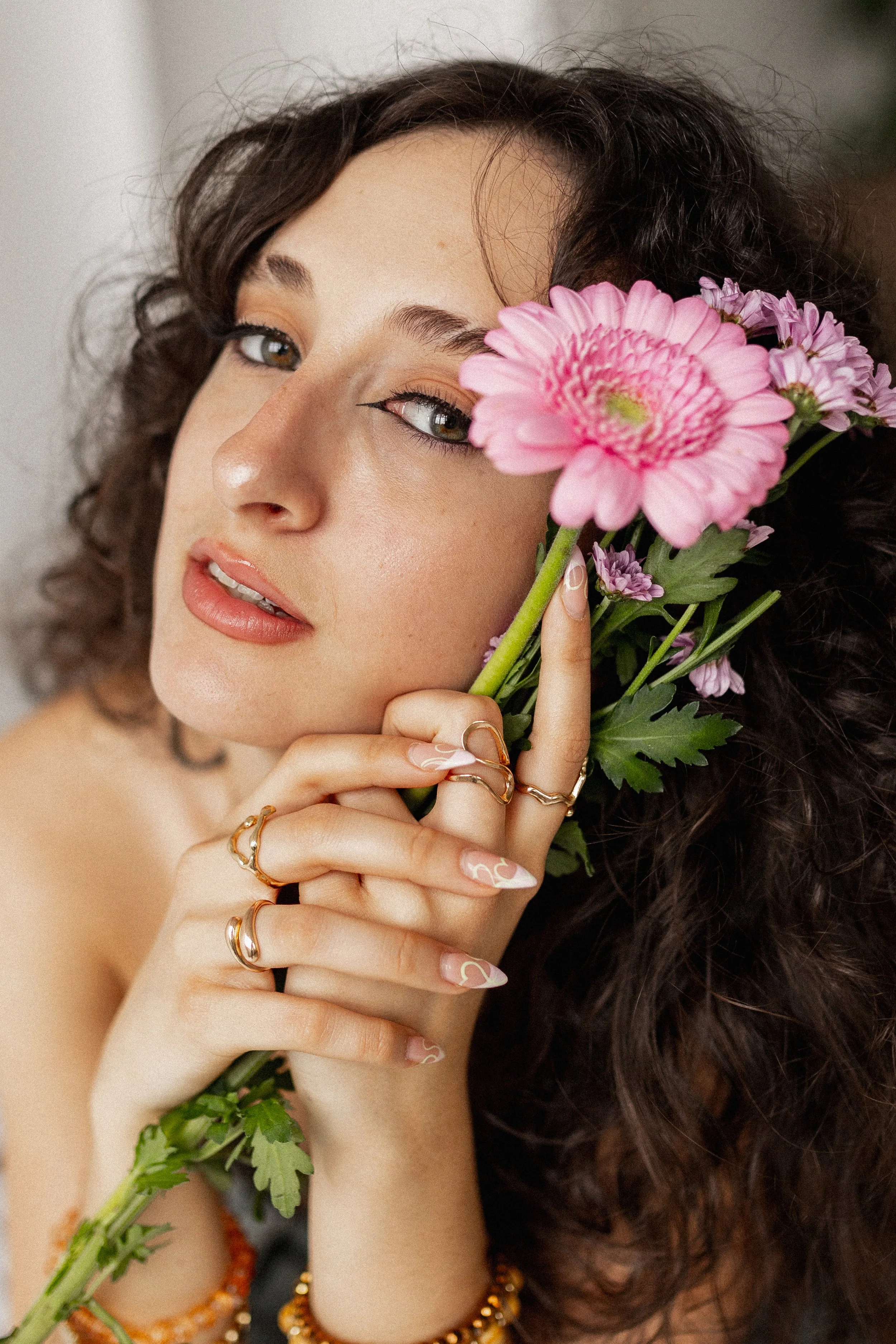A woman with curly dark hair holding pink and purple flowers close to her face, wearing multiple rings and bracelets, with subtle makeup and glossy lips.
