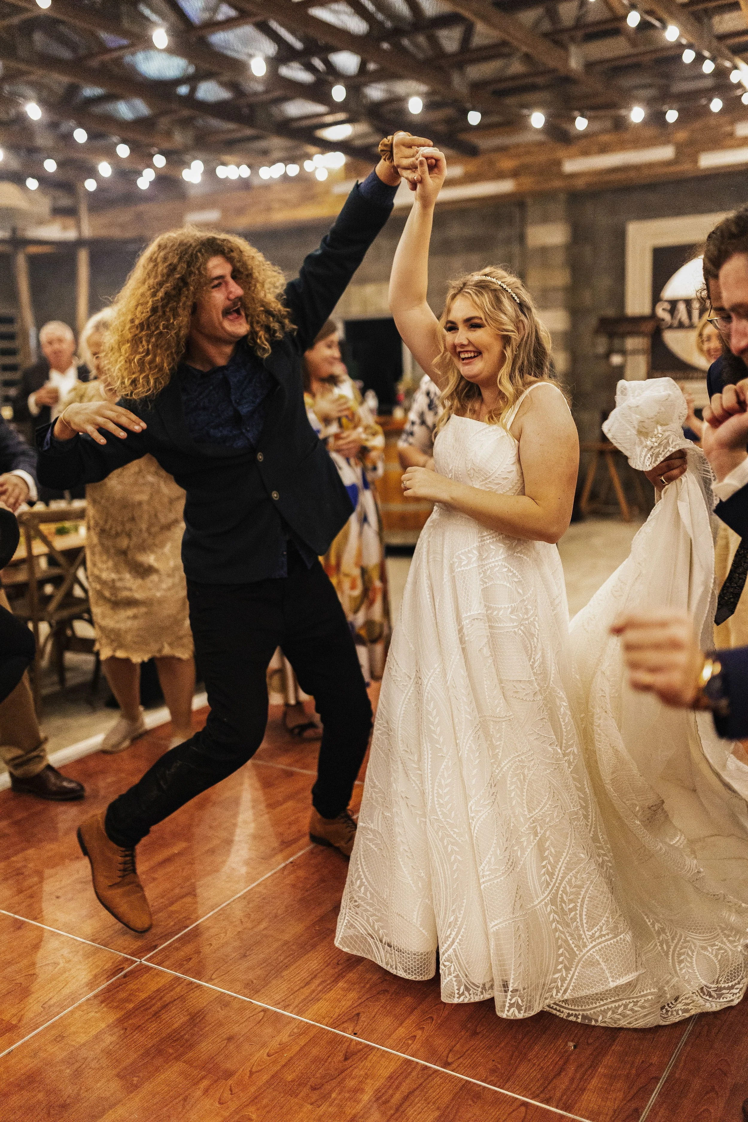 A man and woman dancing happily at a wedding reception, with the man jumping and the woman smiling as they hold hands, surrounded by other guests.