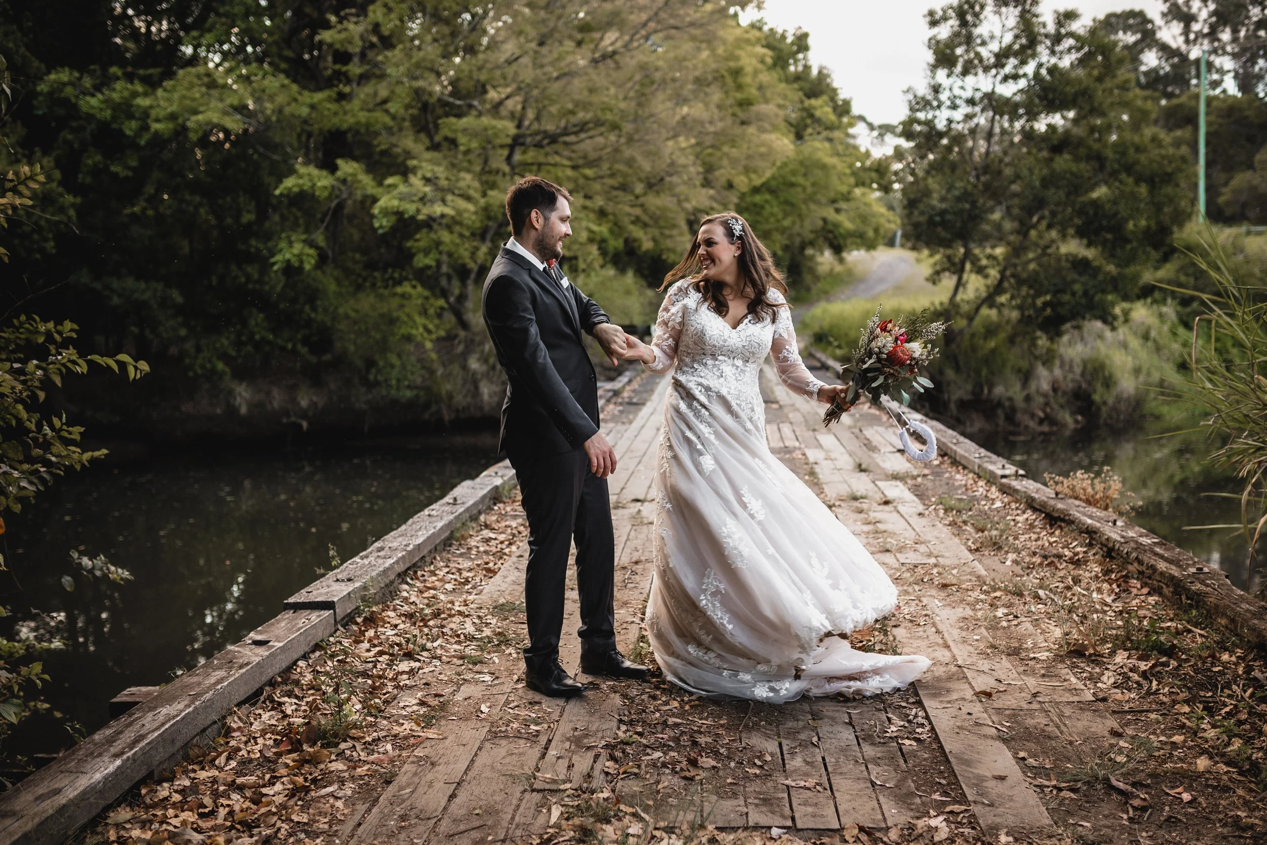 Bride and groom holding hands on a wooden bridge in a lush, green park, with the bride in a white wedding dress and holding a bouquet of flowers.