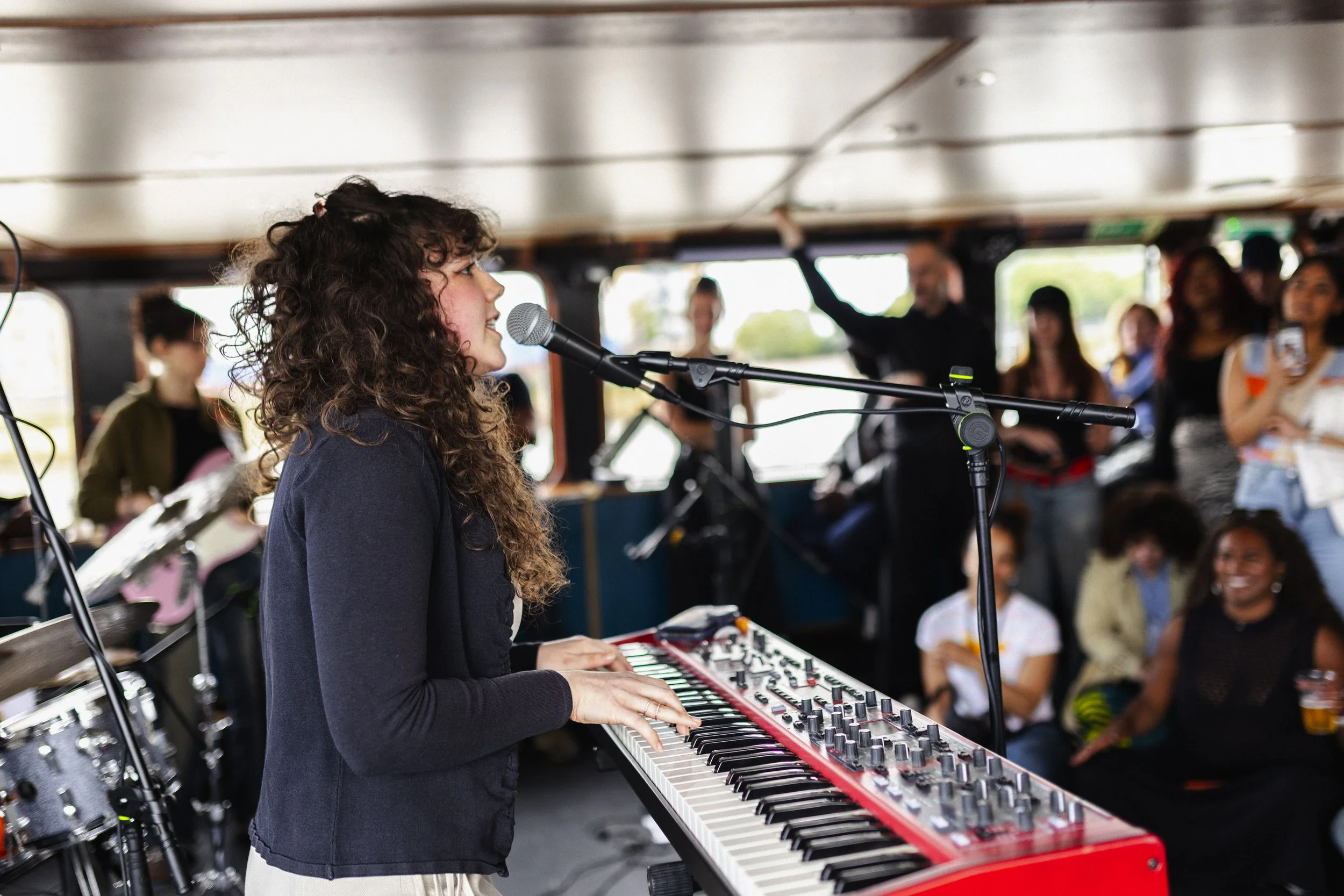 A young woman with curly hair singing into a microphone while playing an electronic keyboard during a live music event on a boat, with an audience in the background.