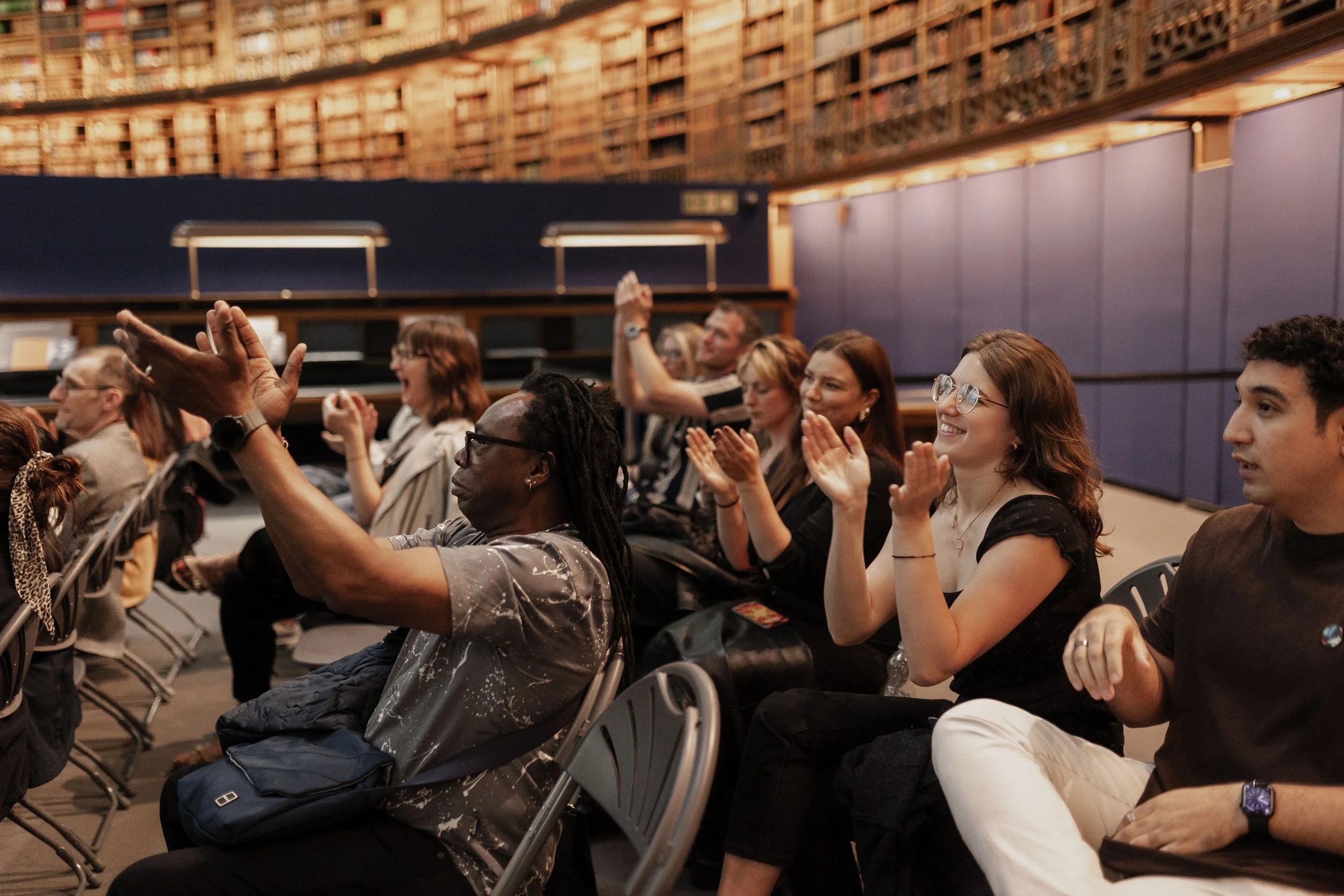 Audience members sitting and smiling during a presentation or event in a large room with bookshelves on the walls.