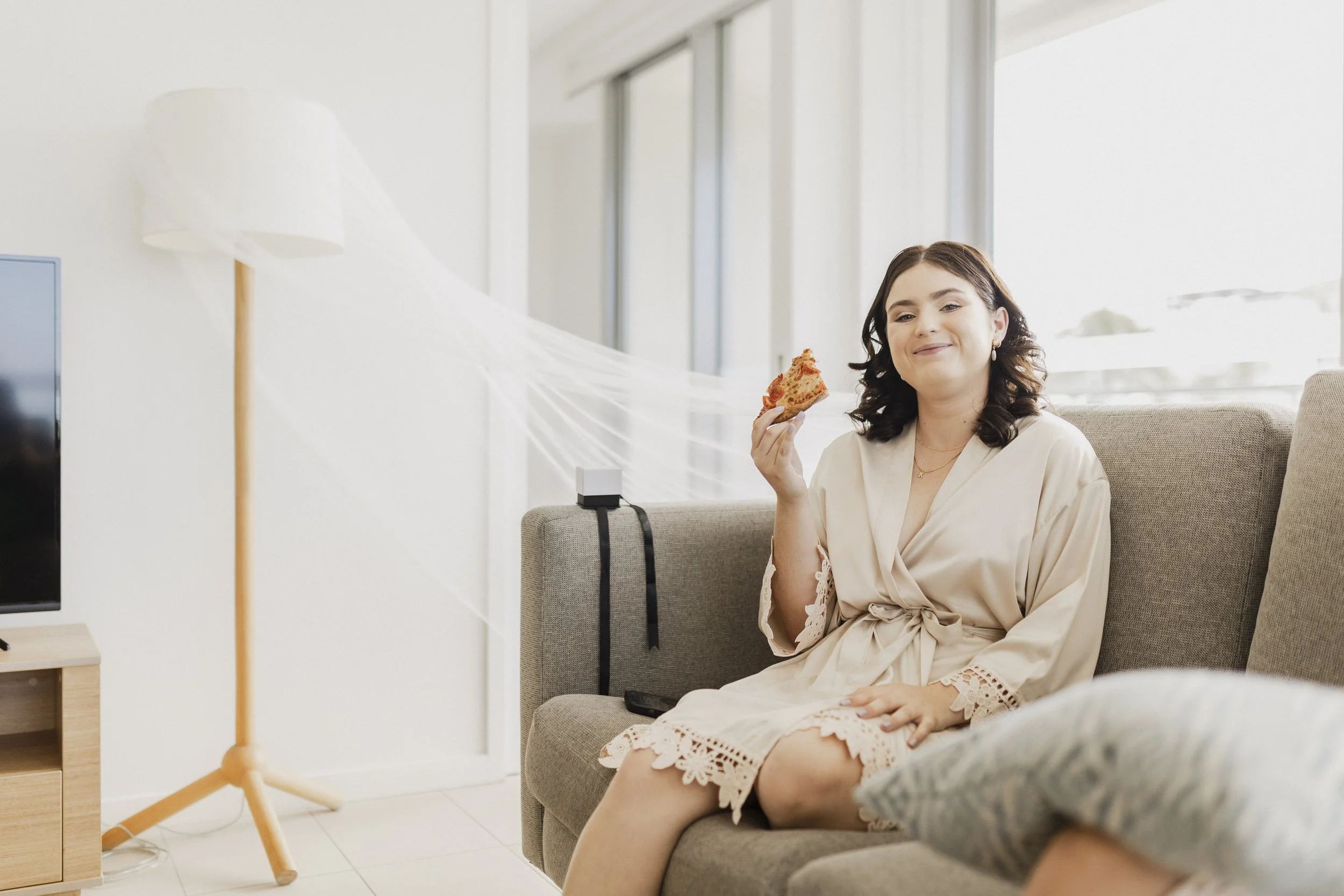 A woman in a beige robe sitting on a gray couch, holding a slice of pizza and smiling at the camera in a brightly lit living room.