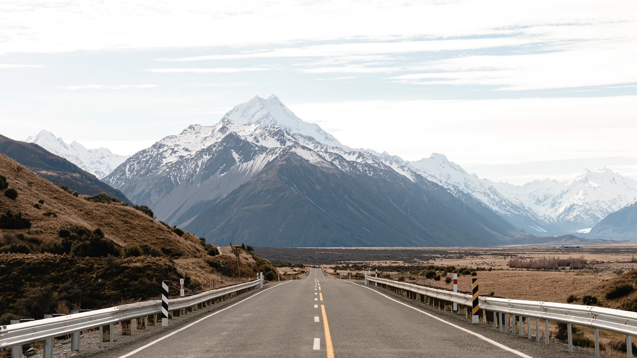 Empty two-lane road stretching into mountainous landscape with snow-capped peaks and dry plains on either side.