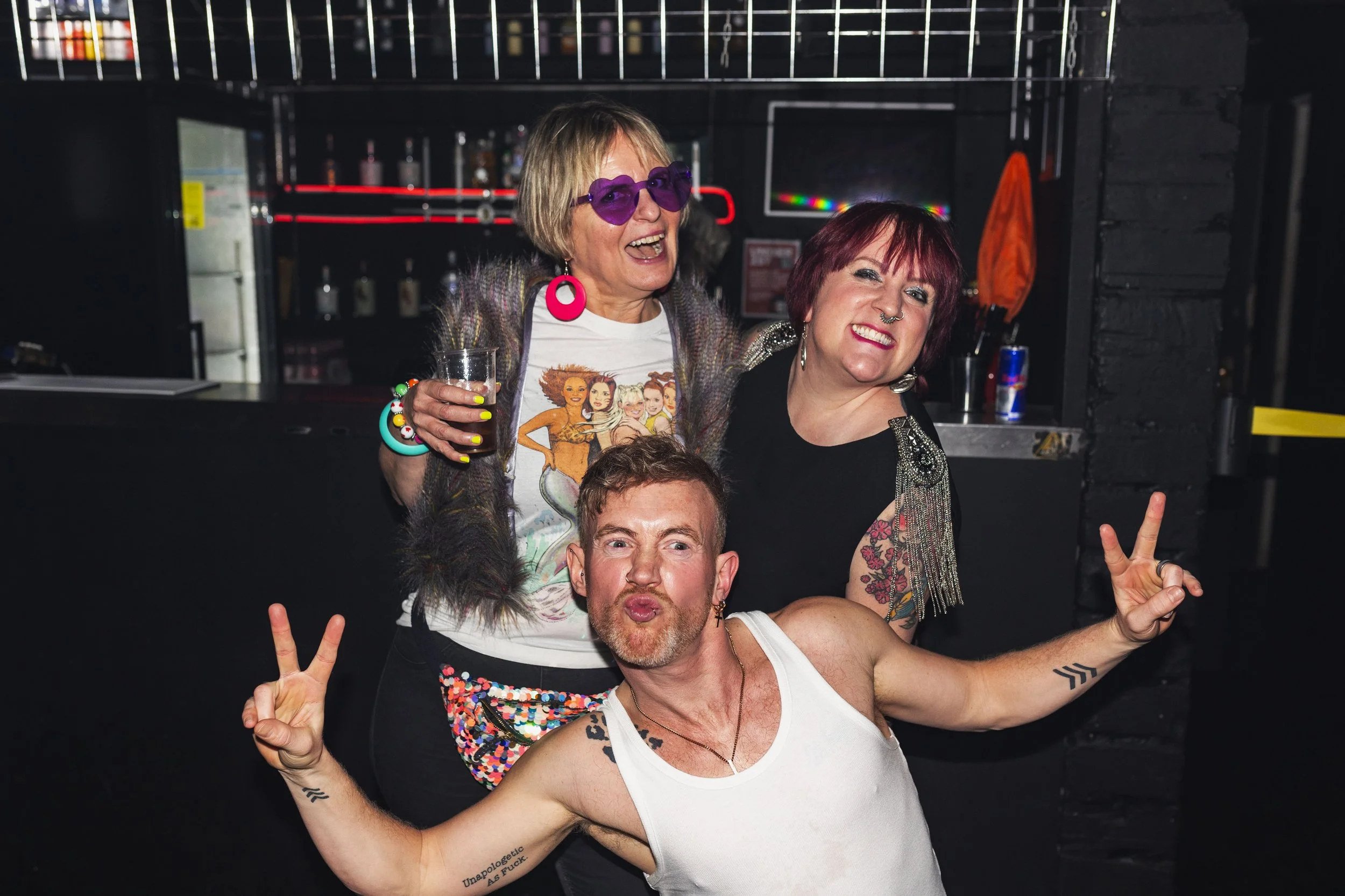 Three people having fun at a bar or nightclub, smiling, making peace signs, with colorful outfits and accessories.