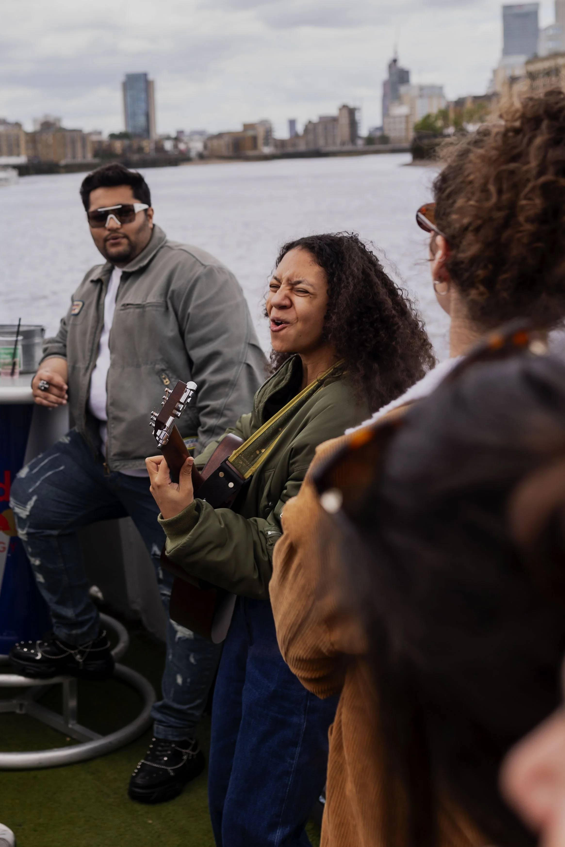 A woman with curly hair singing and playing guitar on a boat with an urban city skyline in the background.