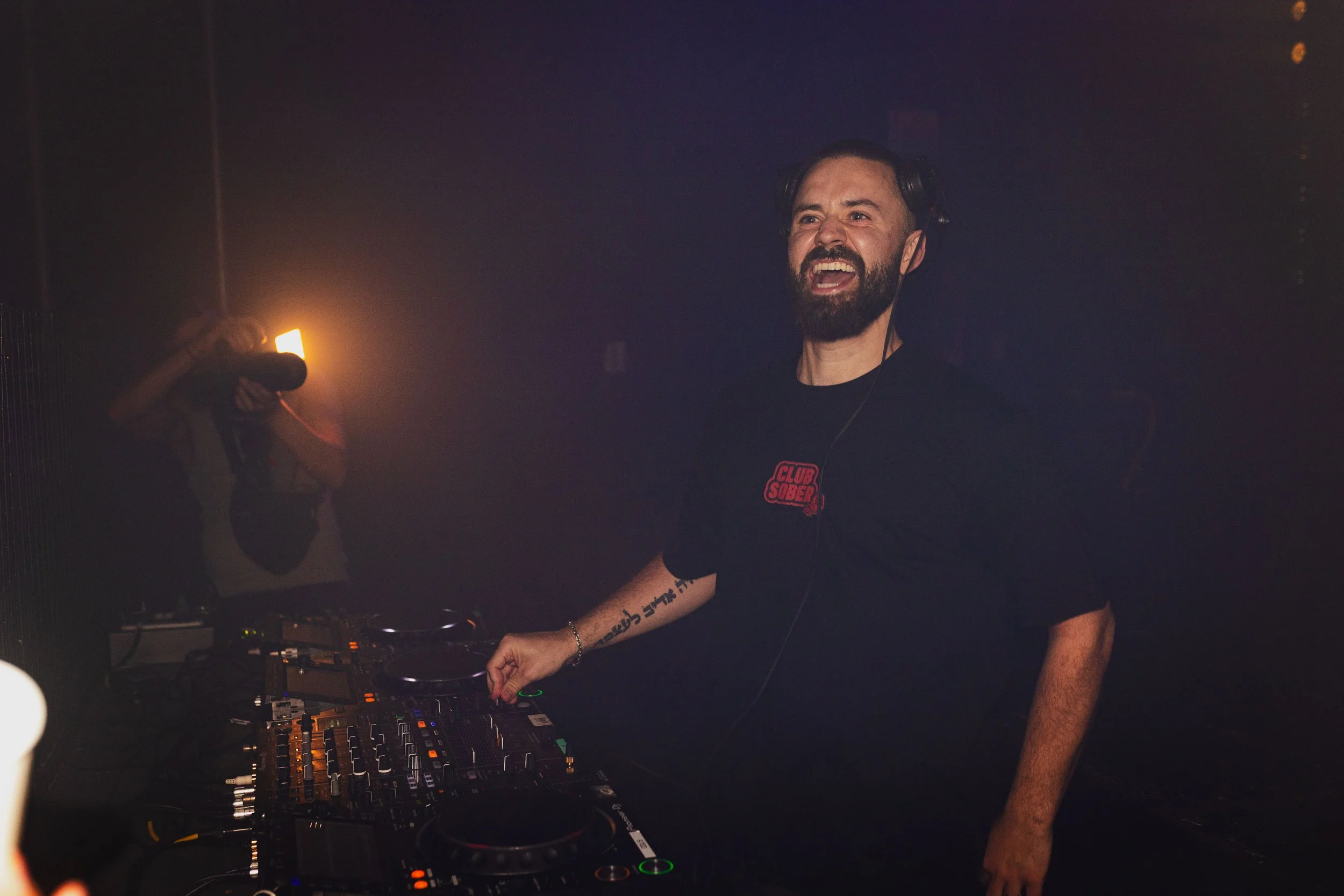 A bearded male DJ wearing headphones, smiling, and operating a DJ controller at a nightclub with dim lighting and a smoky atmosphere.