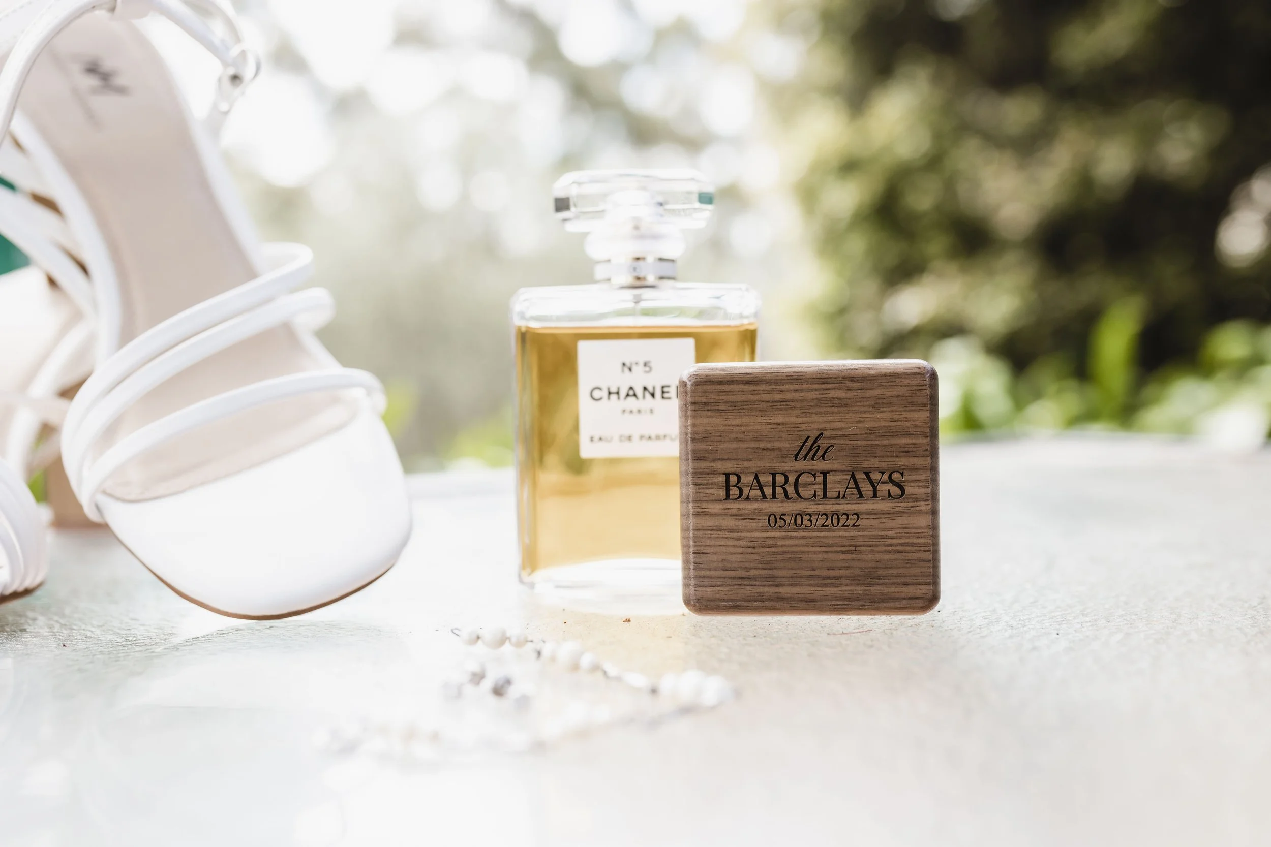 A bottle of Chanel No. 5 perfume, a wooden box engraved with "the BARCLAYS" and a date, white high-heeled shoes with straps, and a white beaded jewelry piece are displayed on a light-colored surface, outdoors with a blurred green background.