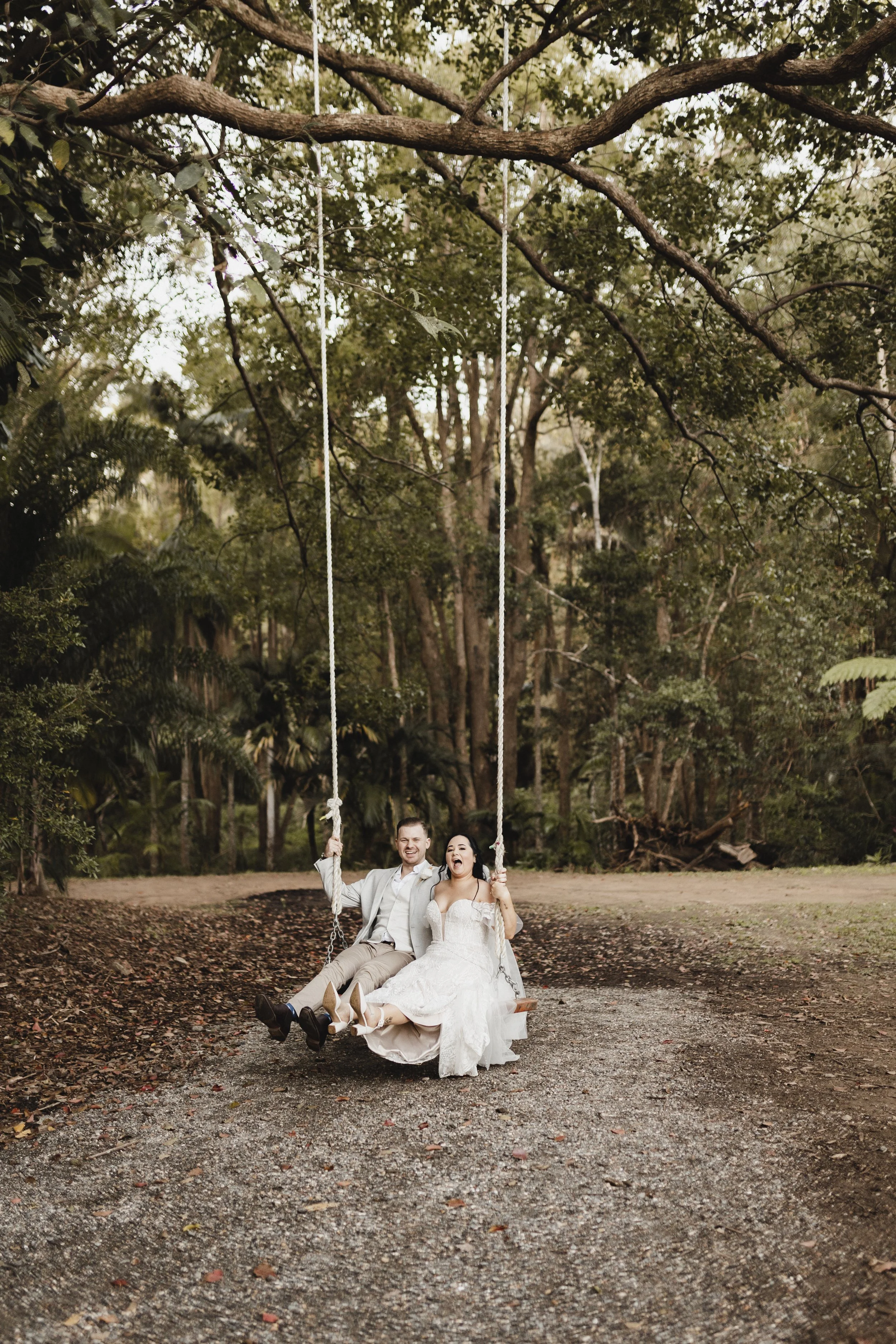 A newlywed couple in formal attire sitting on a wooden swing hanging from a large tree, smiling and enjoying each other's company outdoors in a wooded setting.