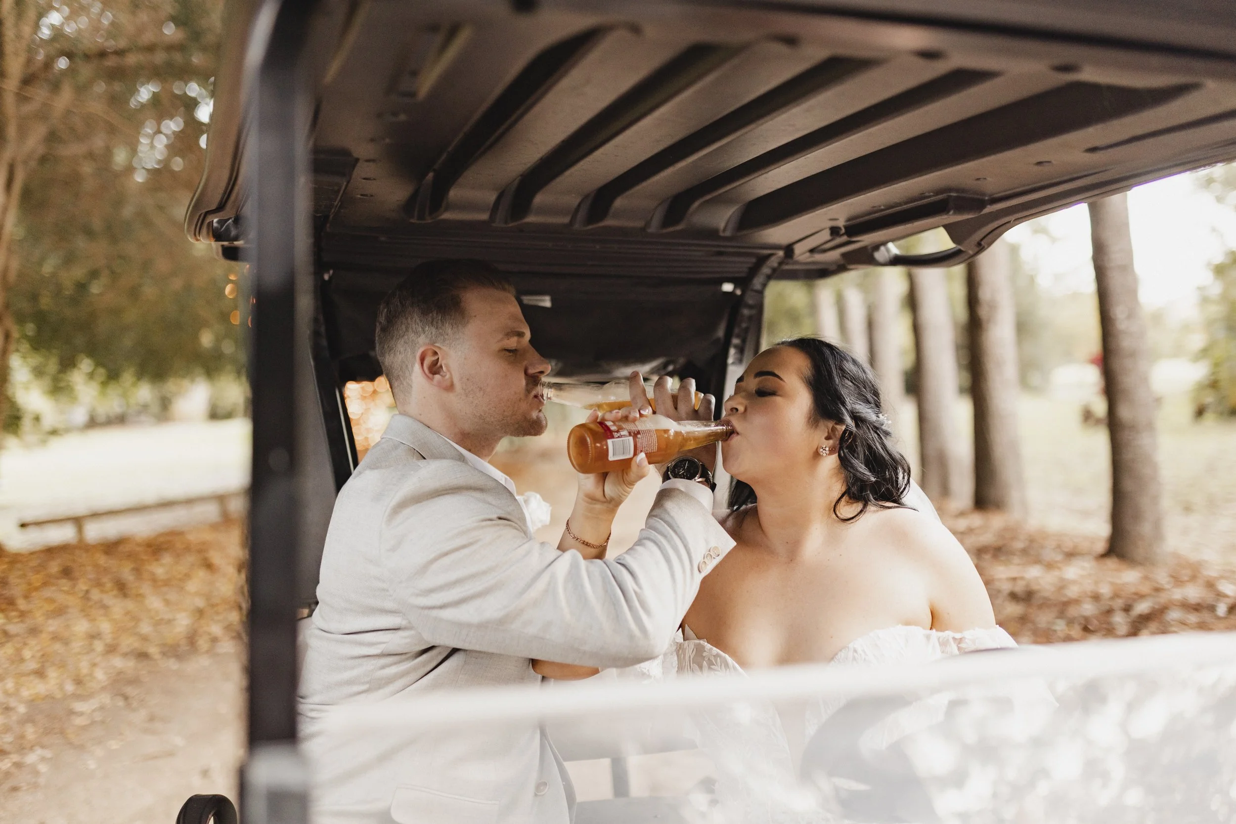A man and woman in wedding attire sharing a toast with bottled beverages inside a golf cart in an outdoor, wooded setting.