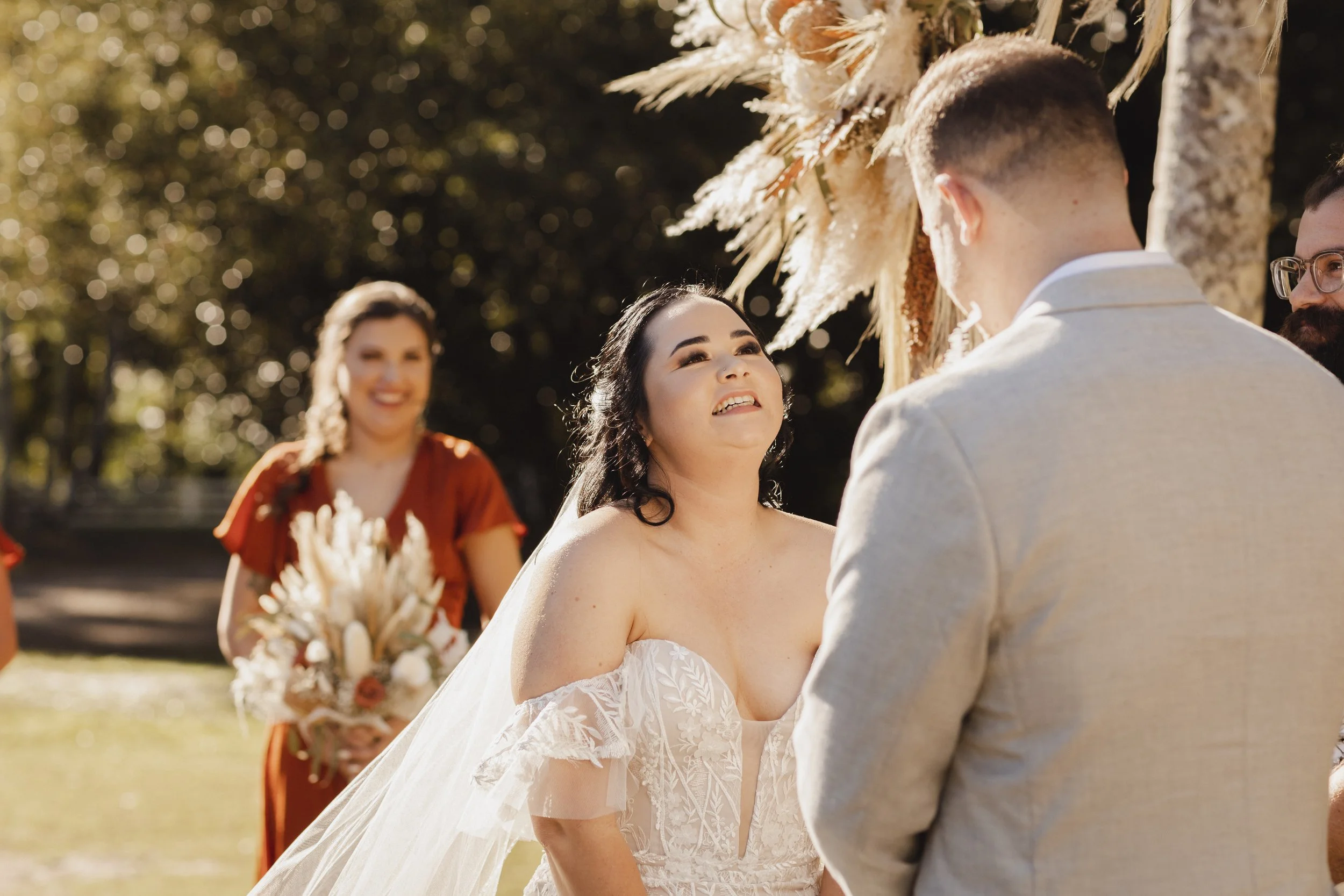 A bride and groom exchange vows outdoors, with a bridesmaid holding a bouquet in the background.