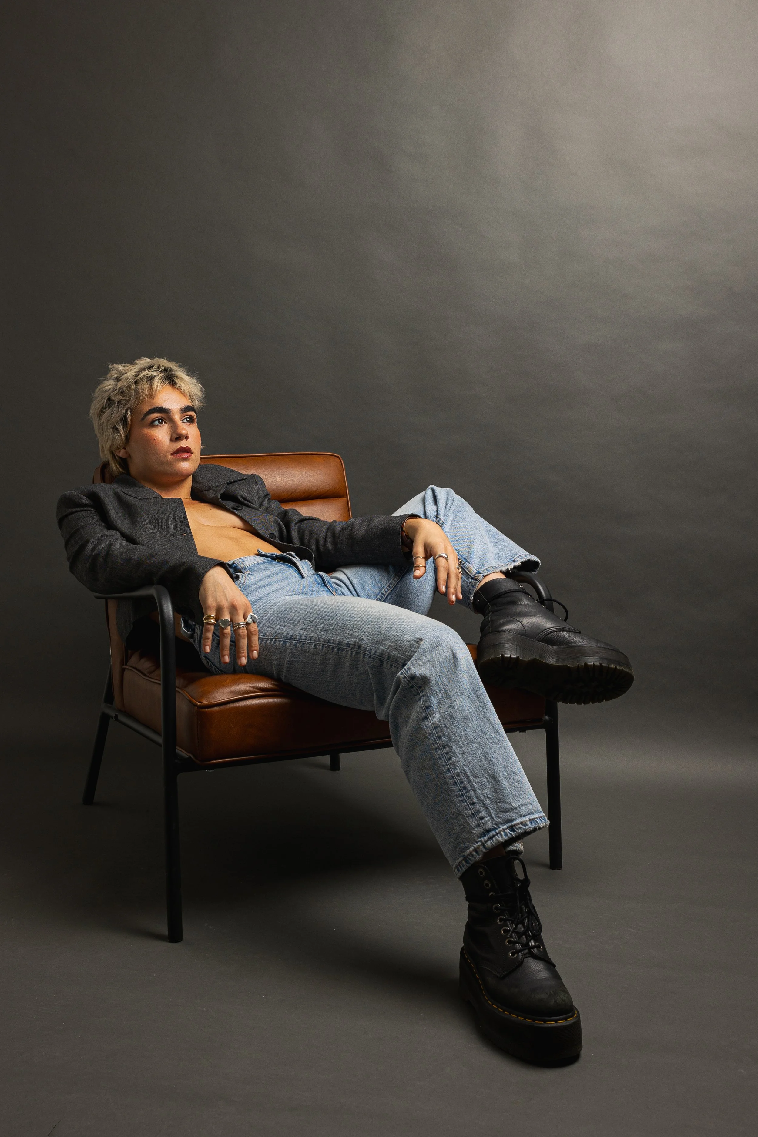 Person with short blonde hair and bold eyebrows sitting on a brown leather chair, wearing a black jacket, blue jeans, and black chunky boots, against a plain gray background.