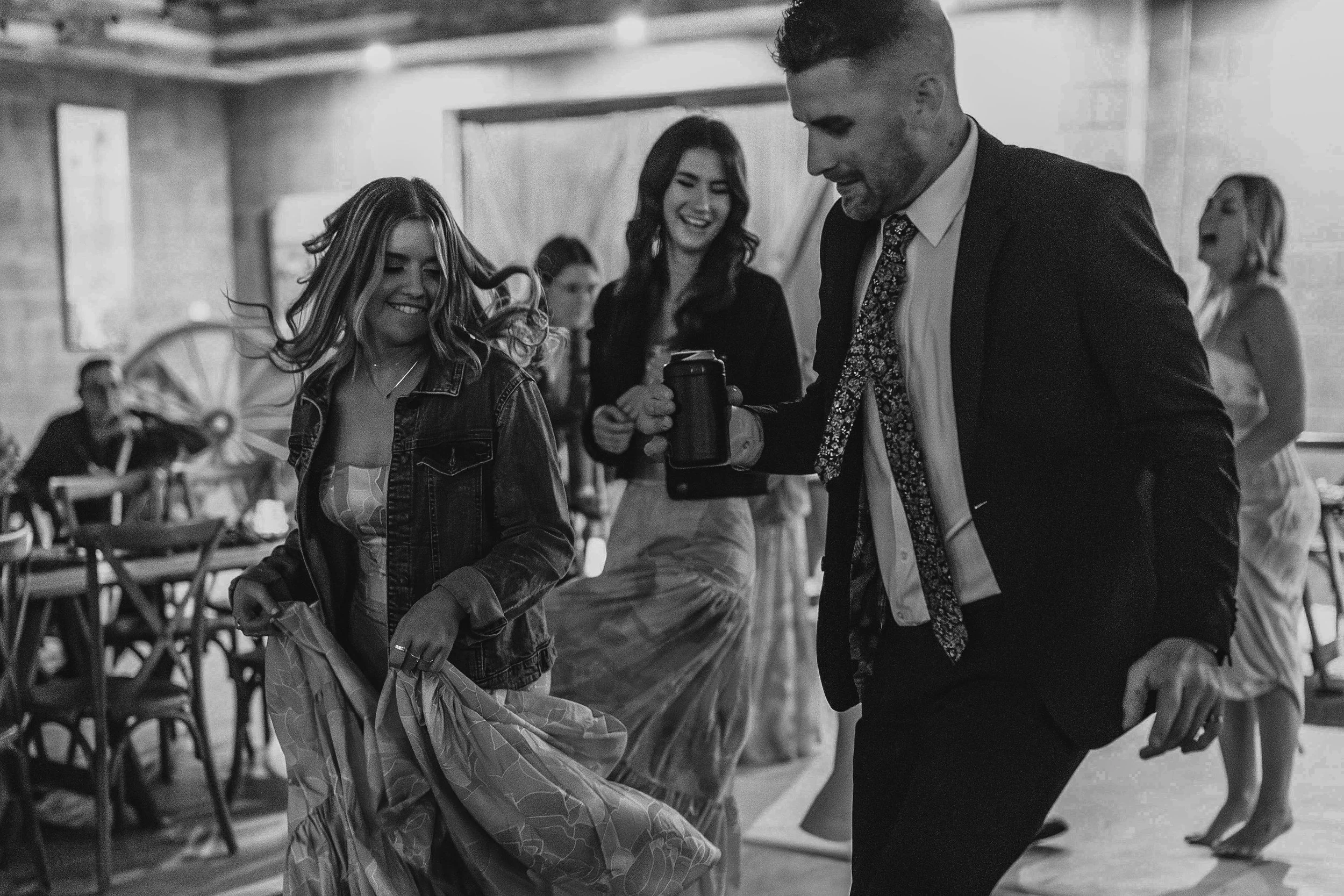 Black and white photo of people dancing at a party, with a young woman in a denim jacket and a man in a suit holding a beverage, enjoying themselves.