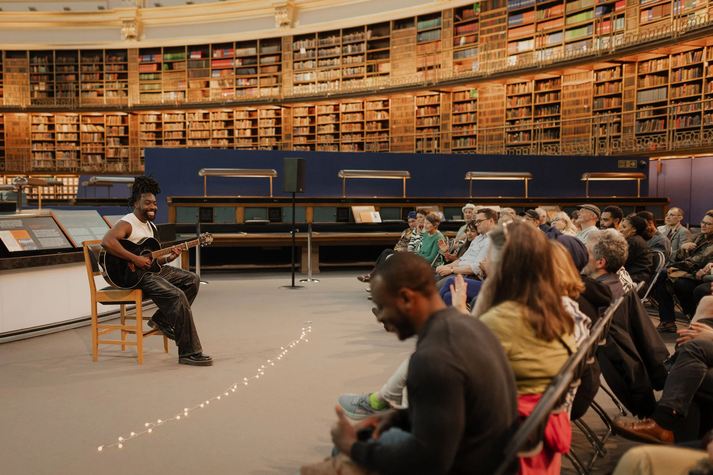A man playing guitar and singing to an audience in a large library or auditorium with bookshelves reaching the ceiling.