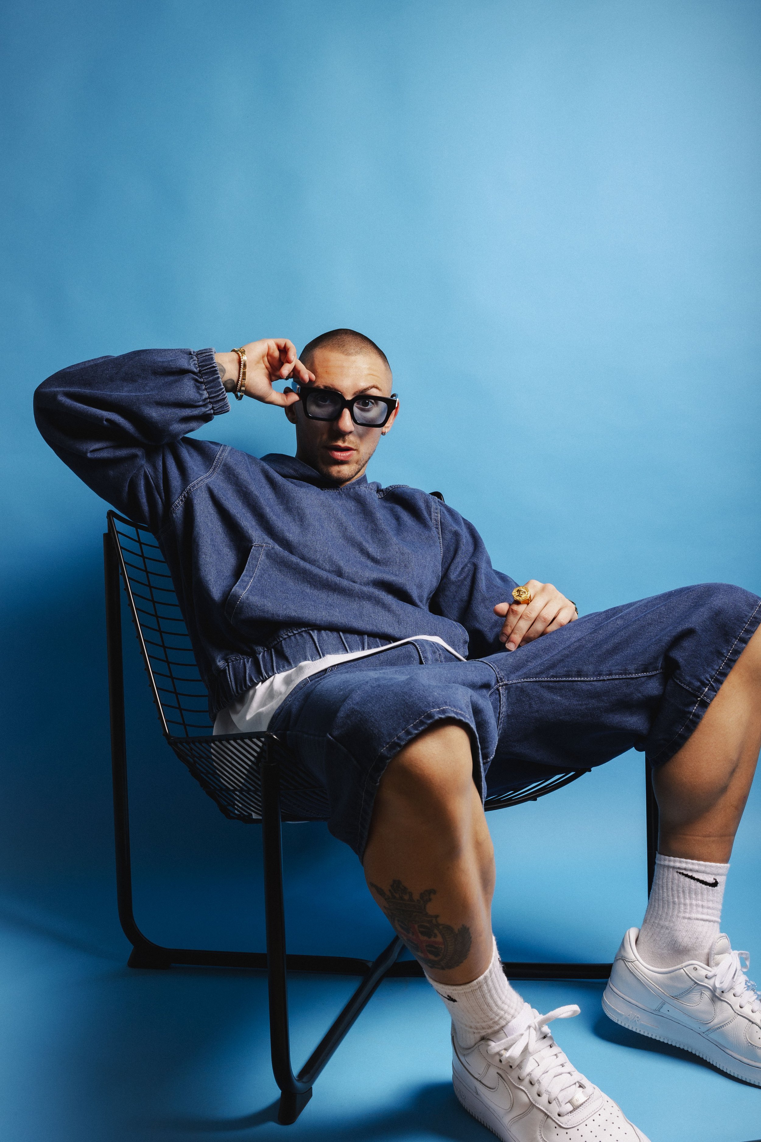 Young man in denim outfit with sunglasses sitting on a wire chair against a blue background