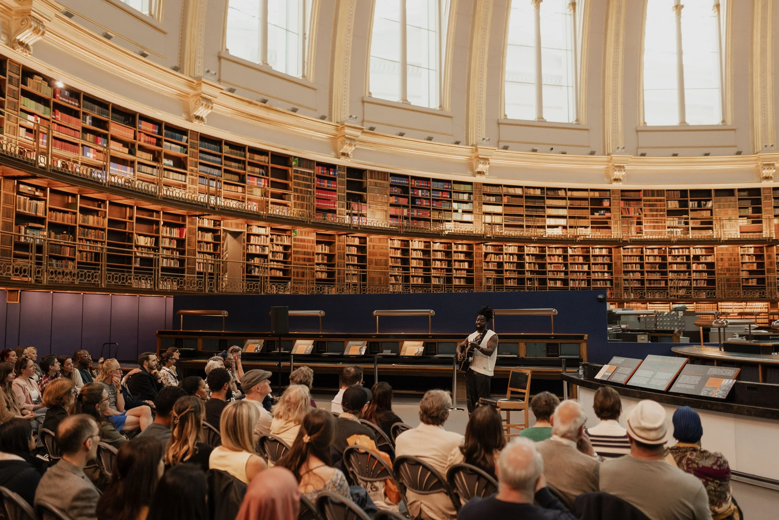 A performer playing guitar in front of an audience in a grand library with tall bookshelves and large windows.