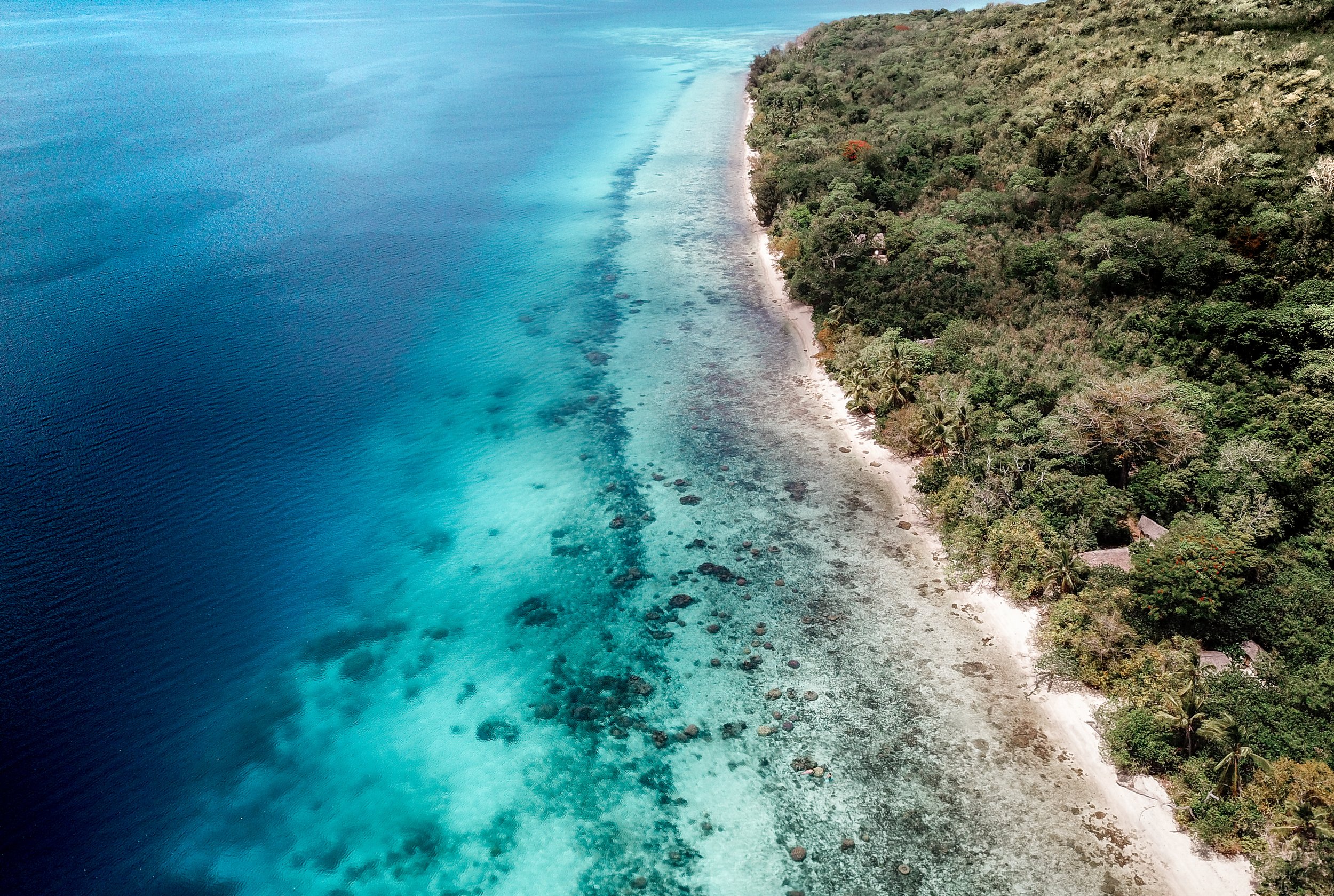 Aerial view of a tropical coastline with clear blue water, coral reefs, white sandy beaches, and dense green forest.