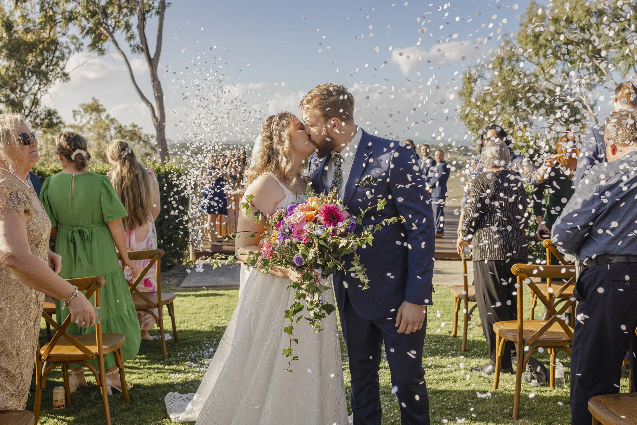 A bride and groom kiss at their outdoor wedding ceremony as guests throw confetti. The bride holds a colorful bouquet; the groom wears a navy suit. Trees and chairs are visible under a sunny sky.
