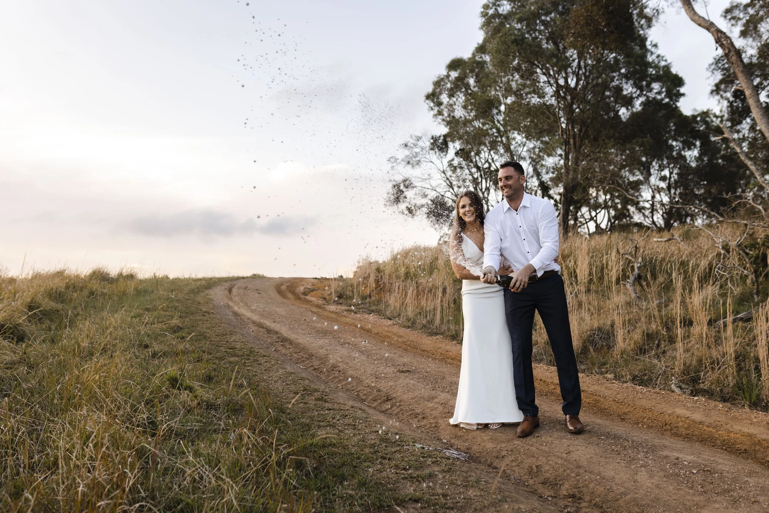 A newlywed couple celebrates outdoors on a dirt road, with the bride in a white dress and the groom in a white shirt and dark pants, popping a champagne bottle as champagne sprays around them, with trees and grass in the background during sunset.
